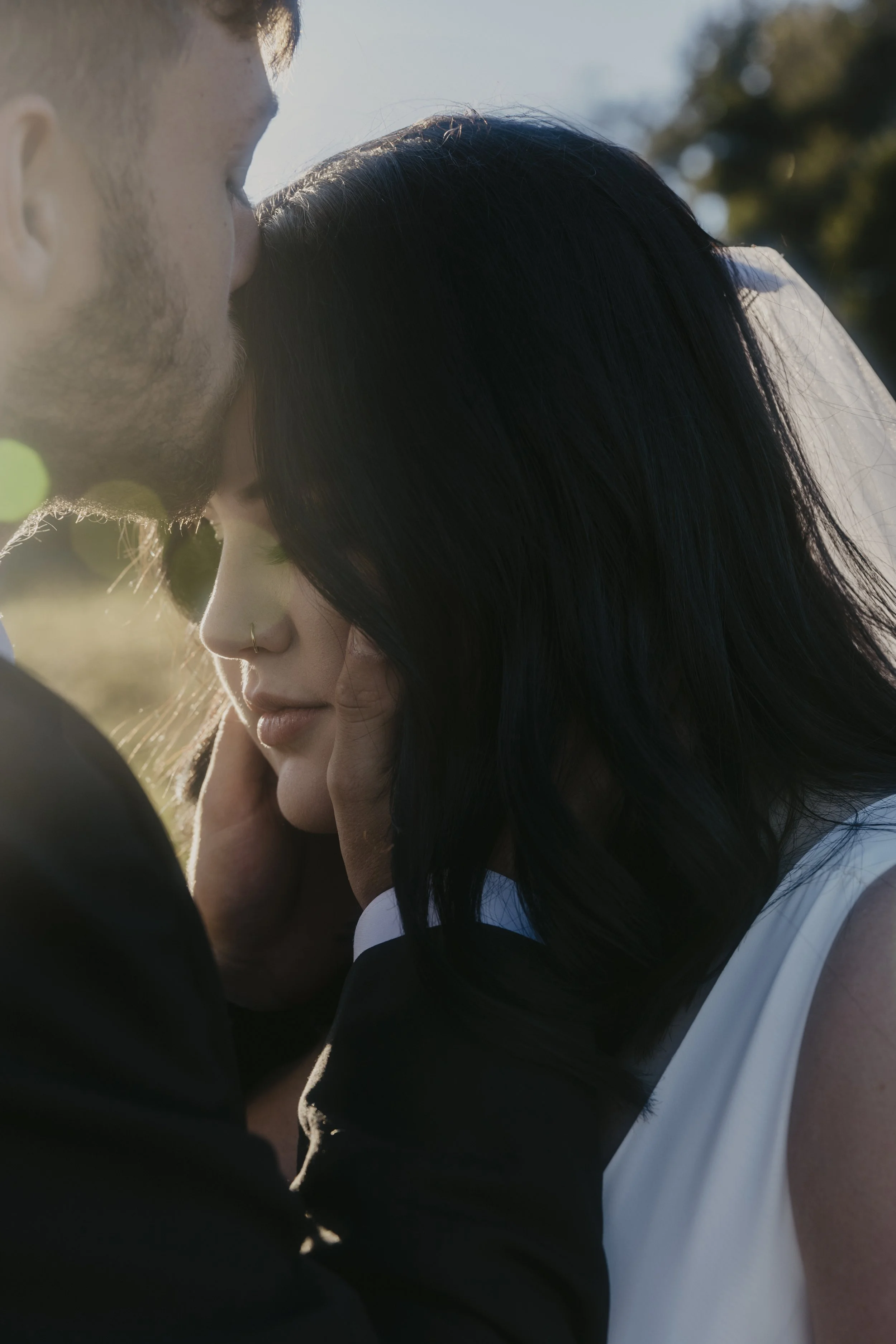 A couple sharing an intimate moment outdoors, with the woman resting her face on their shoulder as the man kisses her forehead, backlit by sunlight.