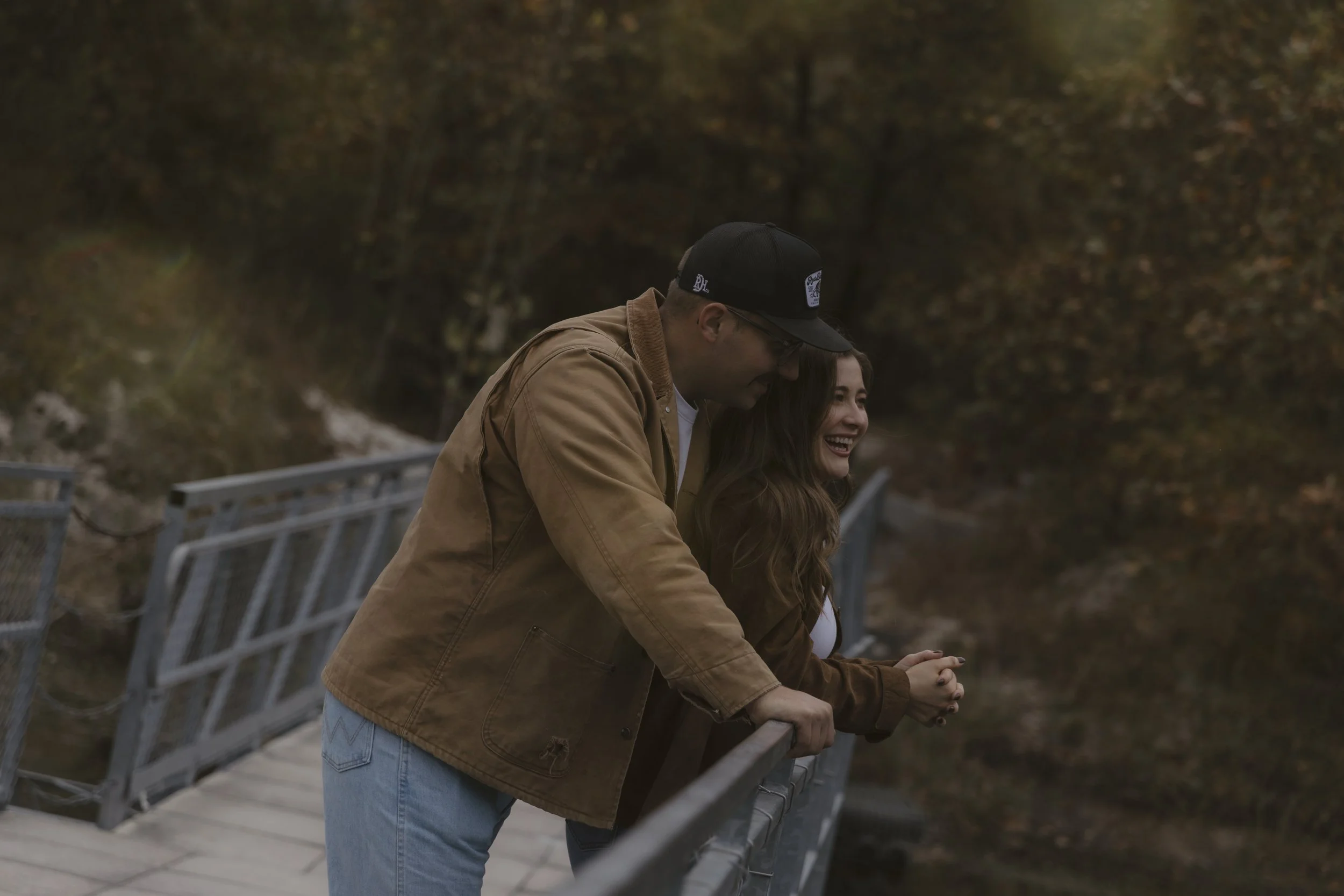 A young man and woman leaning on a bridge railing and looking over a body of water in a forested area during autumn, smiling and enjoying the moment.