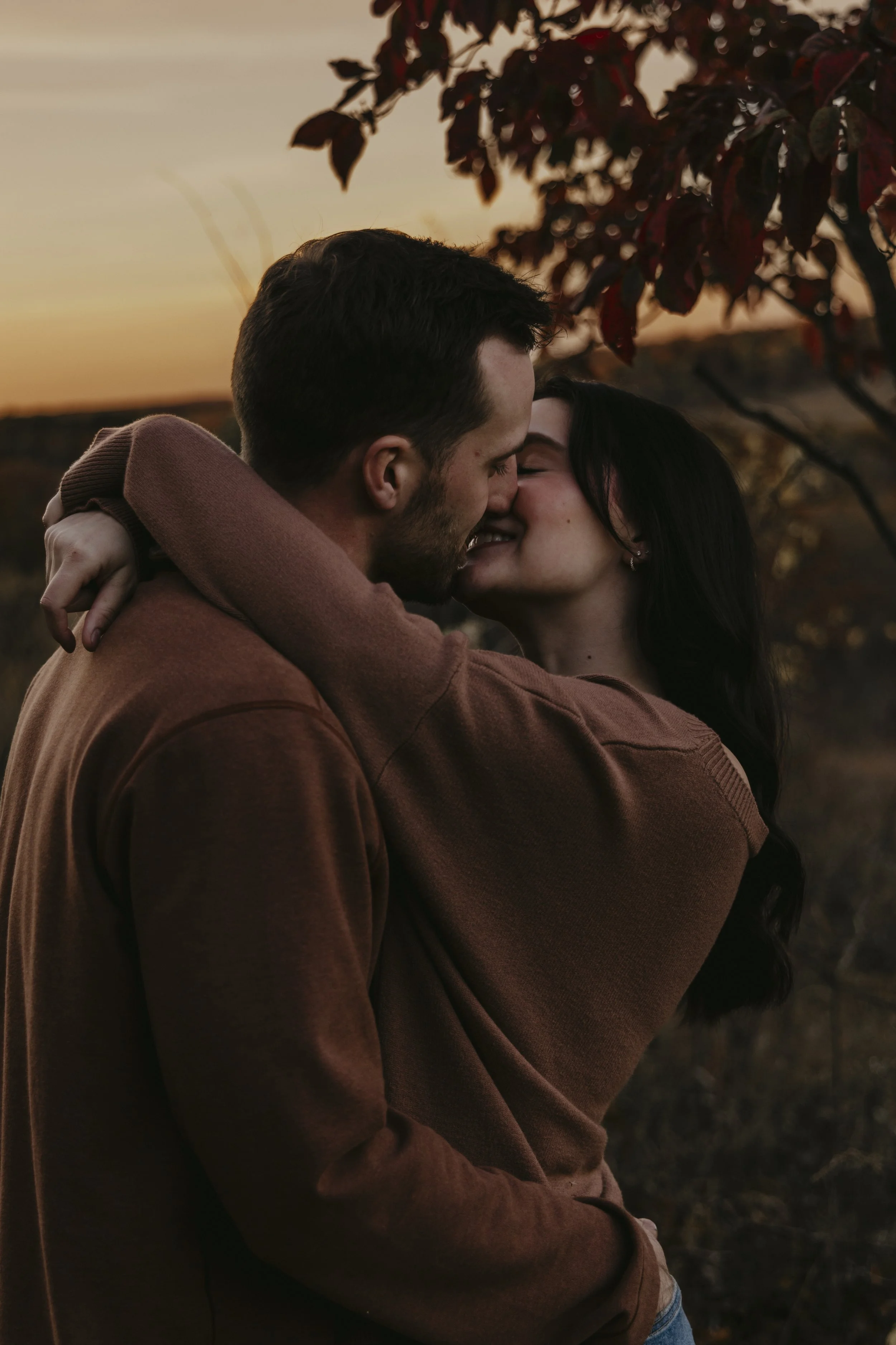 A young couple embracing and kissing outdoors during sunset, with trees and warm orange sky in the background.