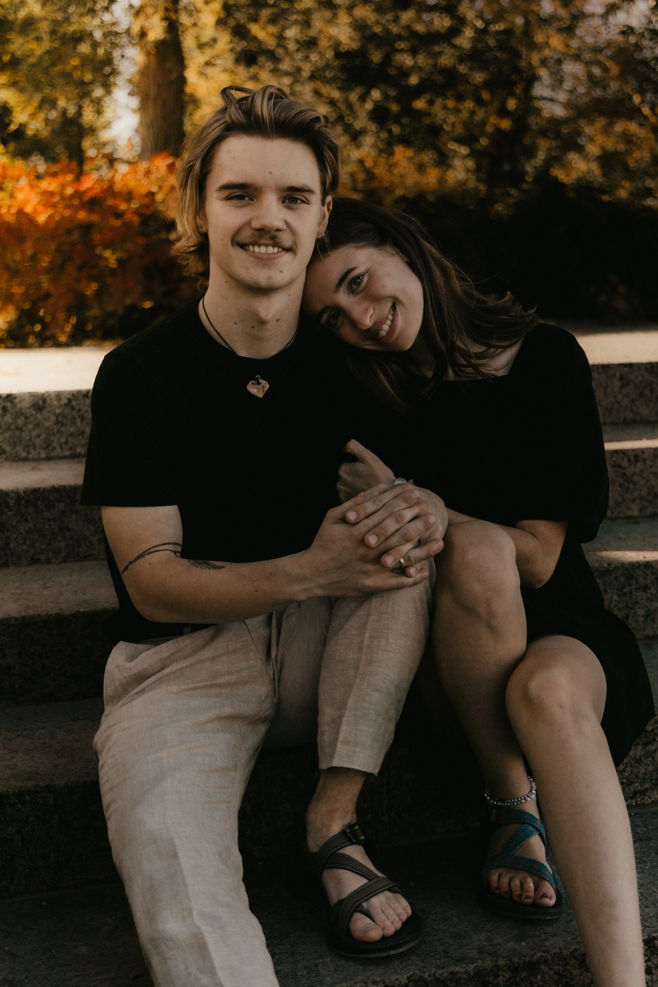 A young couple sitting closely together on outdoor stairs, smiling, with autumn trees and foliage in the background during daytime.