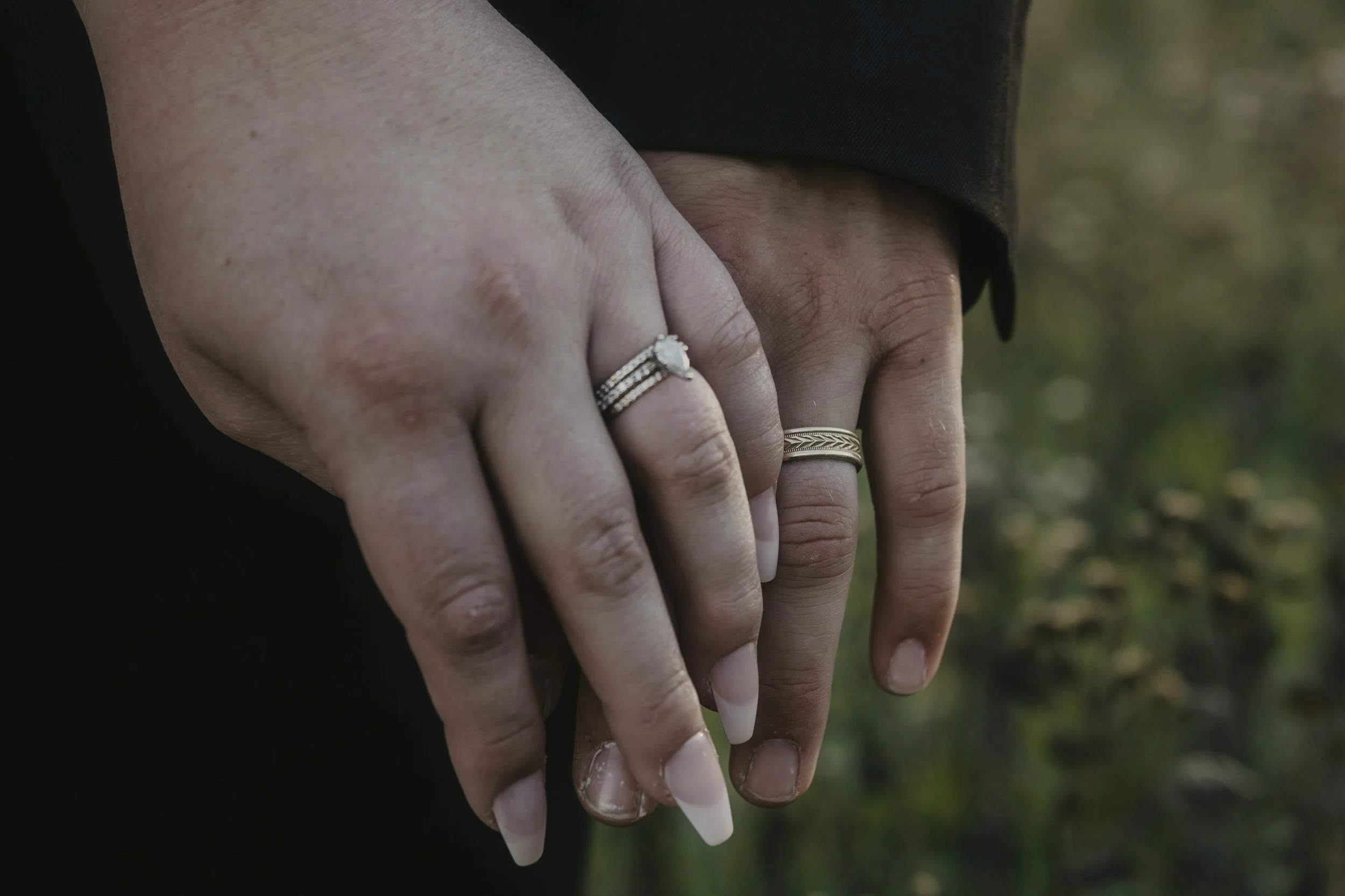 Close-up of two hands holding each other, wearing wedding and engagement rings, with a blurred natural background.