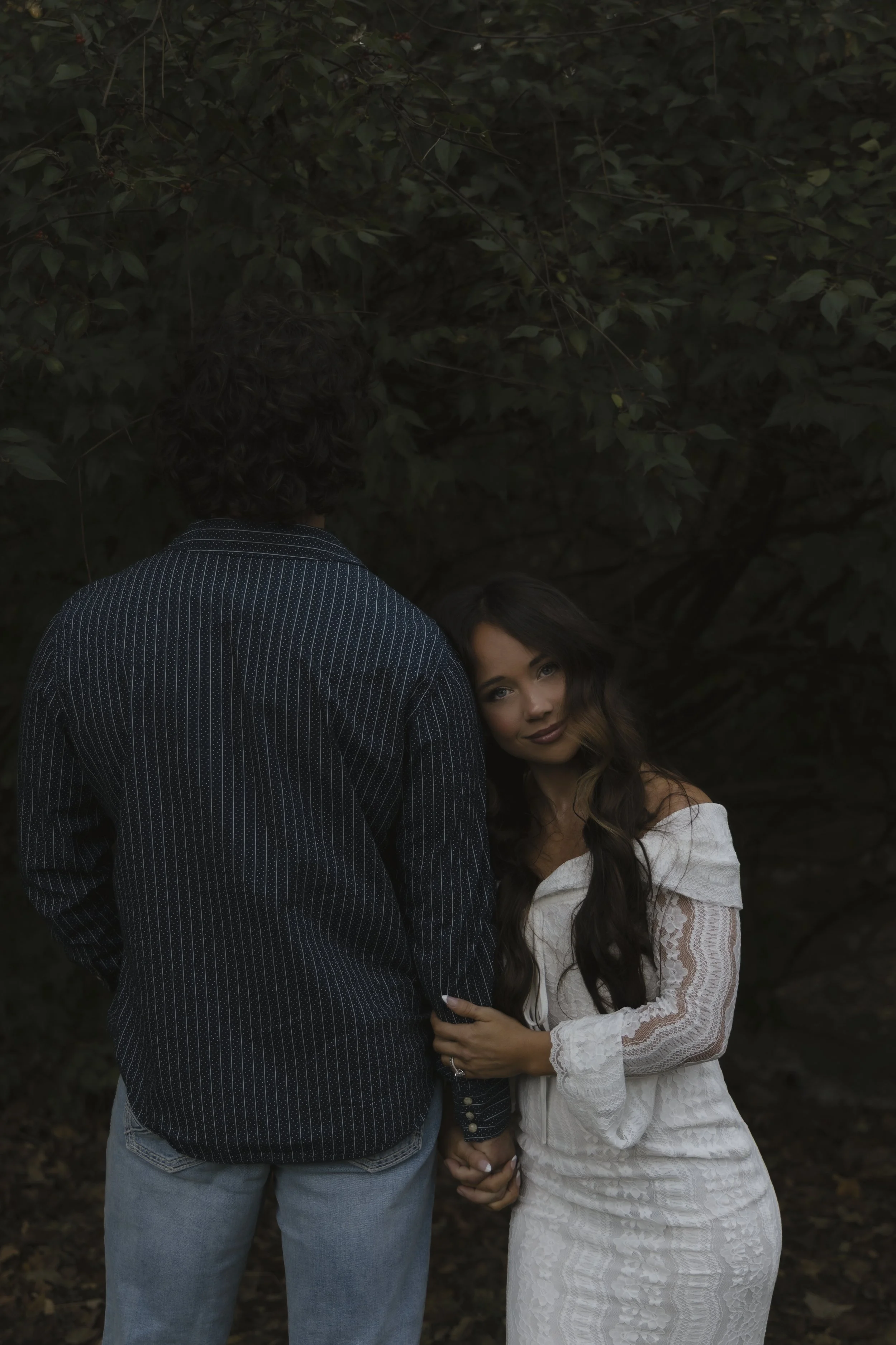 A woman in a white lace off-shoulder dress holding hands with a man in a dark striped shirt and light jeans, standing outdoors amid greenery.