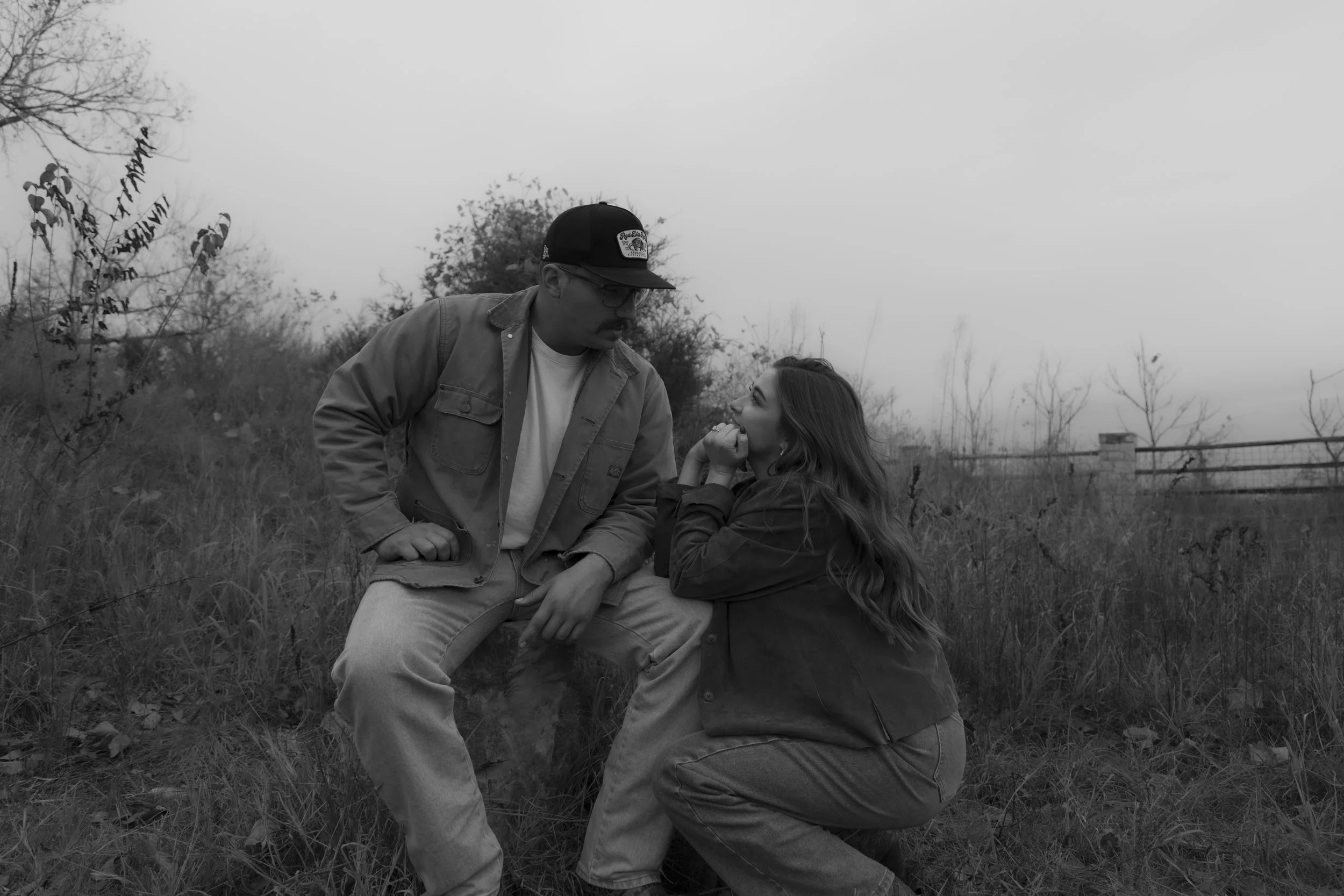 A man and woman sitting on grass outdoors, facing each other closely, with the woman smiling and touching her chin, and the man looking down at her. Tree branches and a fence are visible in the background.