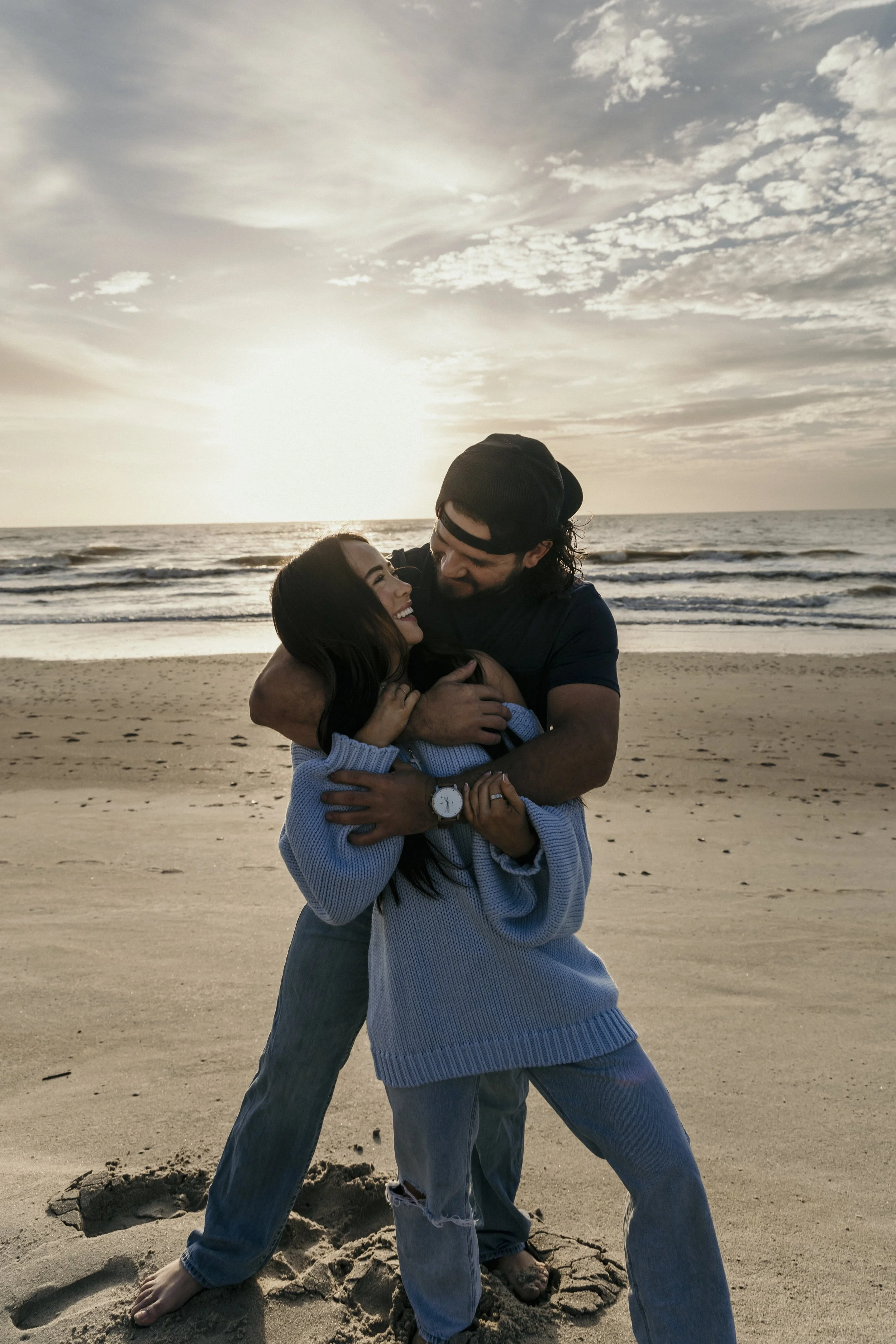 A couple embracing on a beach during sunset, smiling and looking at each other, with the ocean and sky in the background.