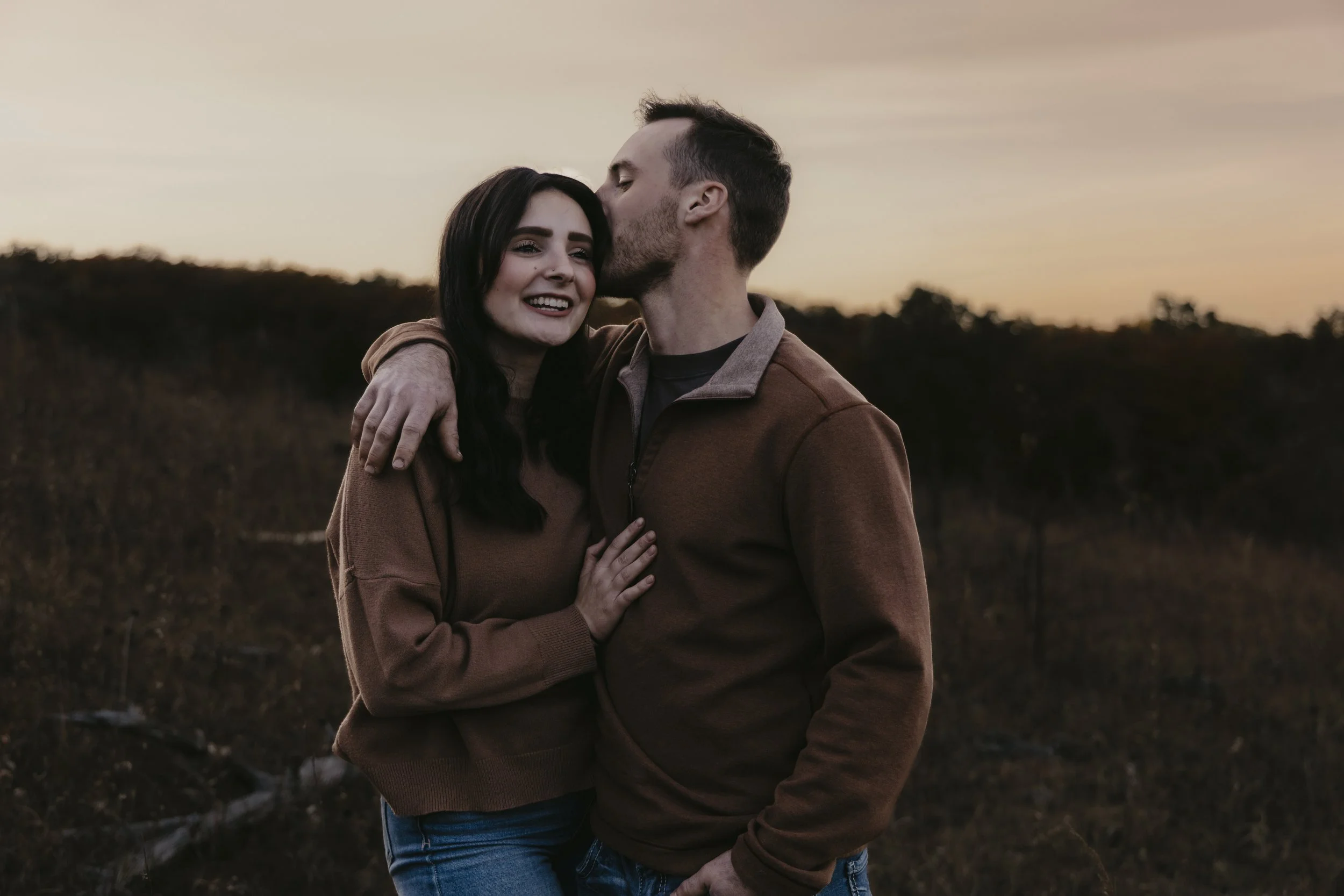 A young couple hugging outdoors at sunset, with the man kissing the woman's temple, both smiling.