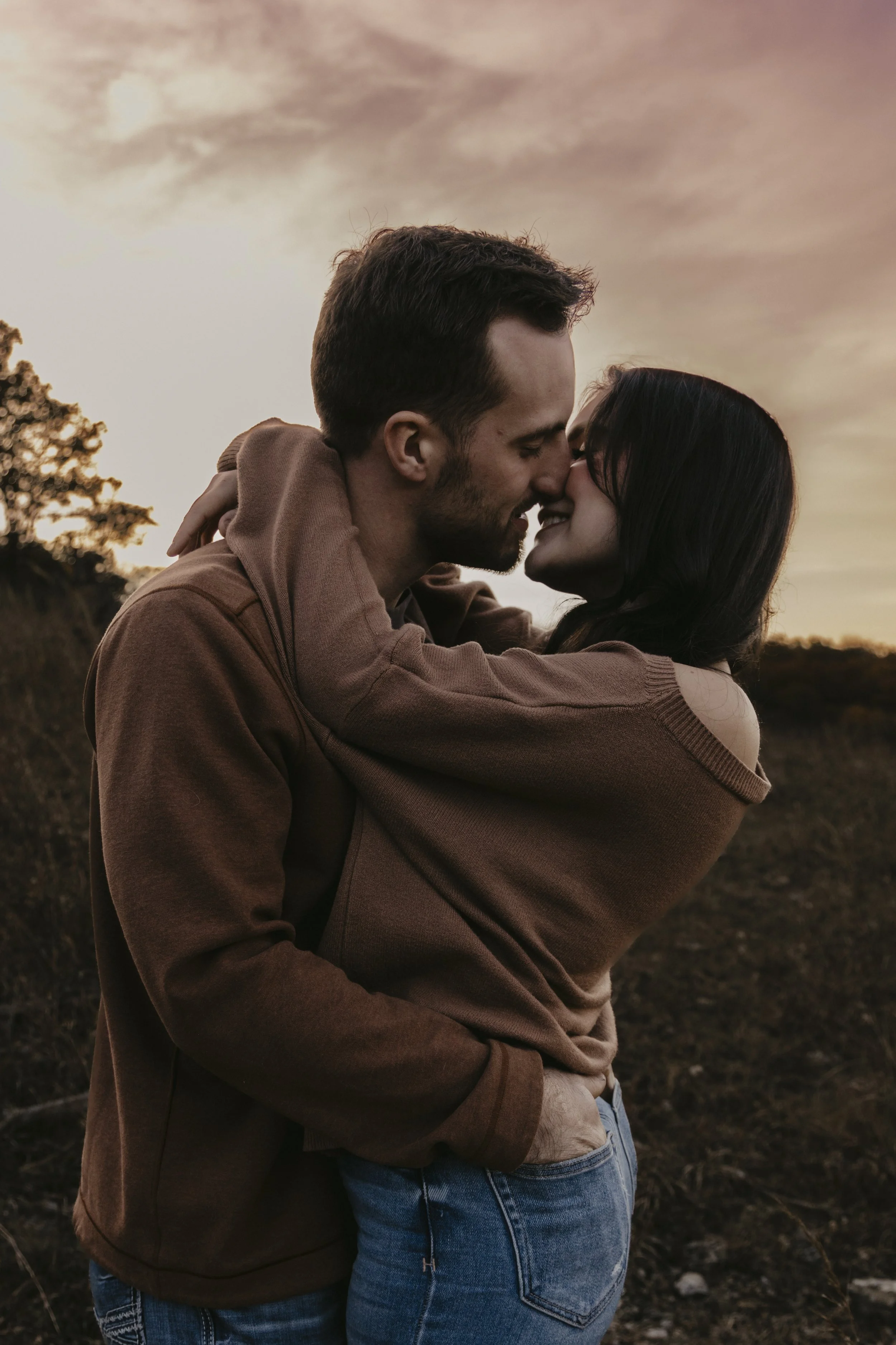 A couple embracing and about to kiss outdoors at sunset, with a cloudy sky and trees in the background.