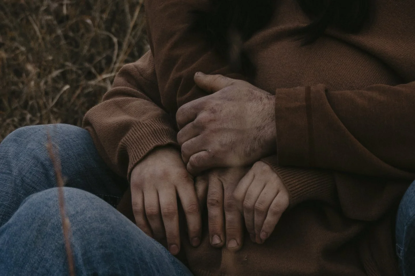 Close-up of two people holding hands, with one person's hand on top of the other's. Both appear to have rough, dirty hands. They are wearing casual clothes, one in a brown sweater and the other in blue jeans. The background shows dry grass.
