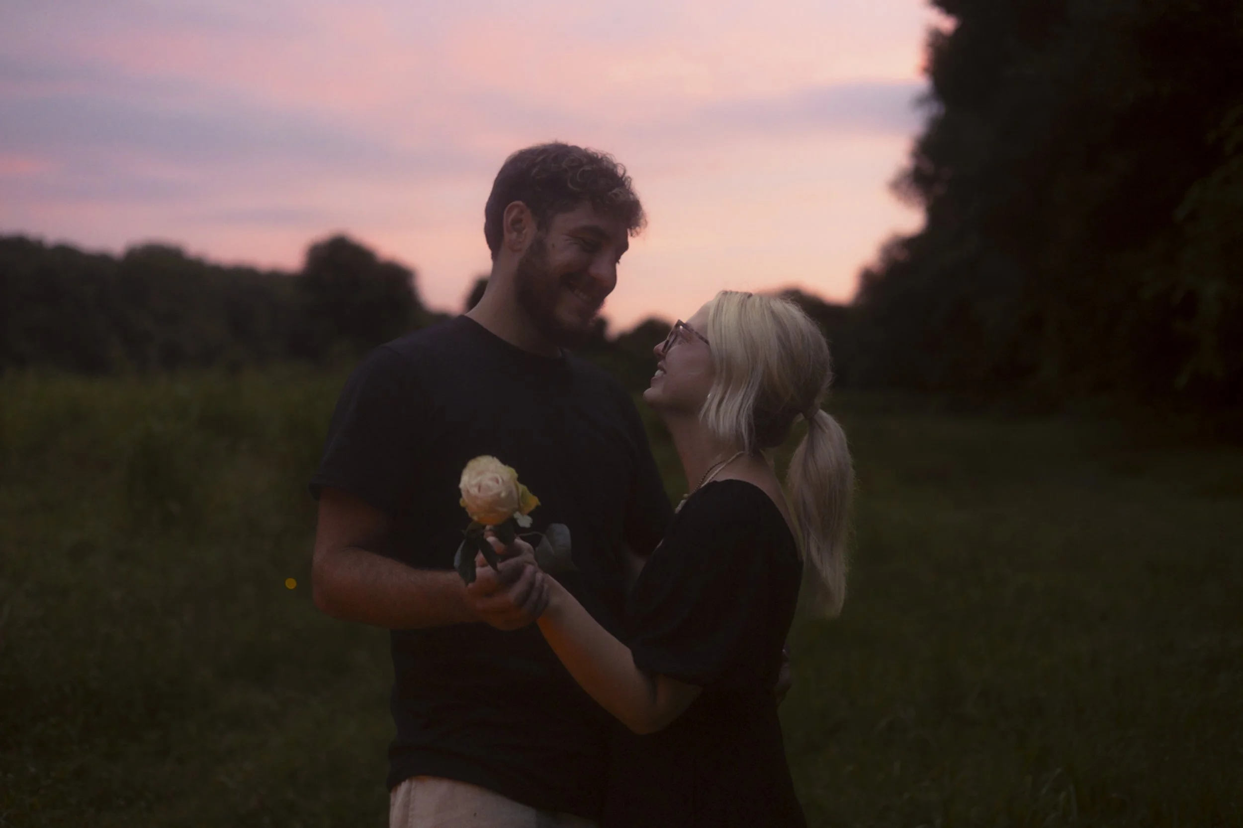 A couple standing close together outdoors during sunset, smiling at each other. The man holds a flower in his hand, and the woman gazes up at him. Trees and a pink and purple sky are in the background.