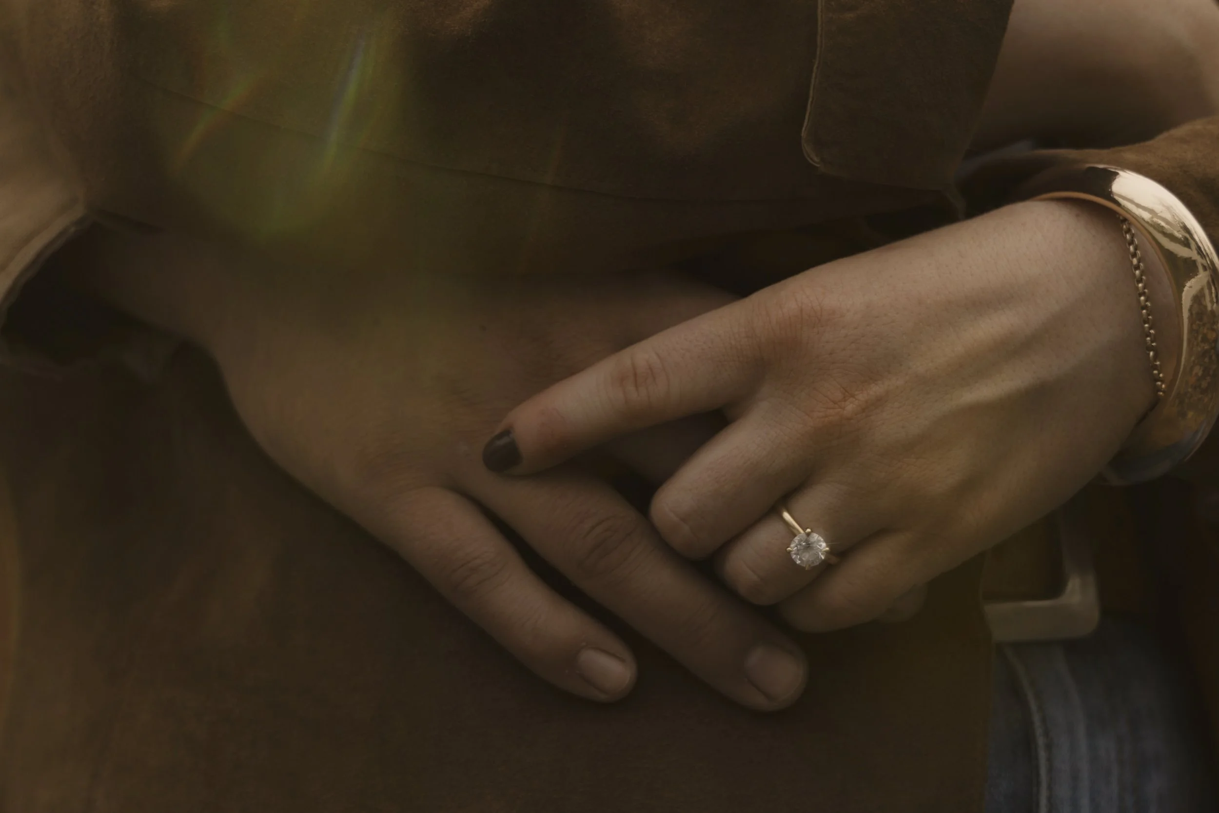 Close-up of a woman's hand with a diamond engagement ring on her ring finger, resting on her lap, wearing a gold bracelet and a wrist chain.