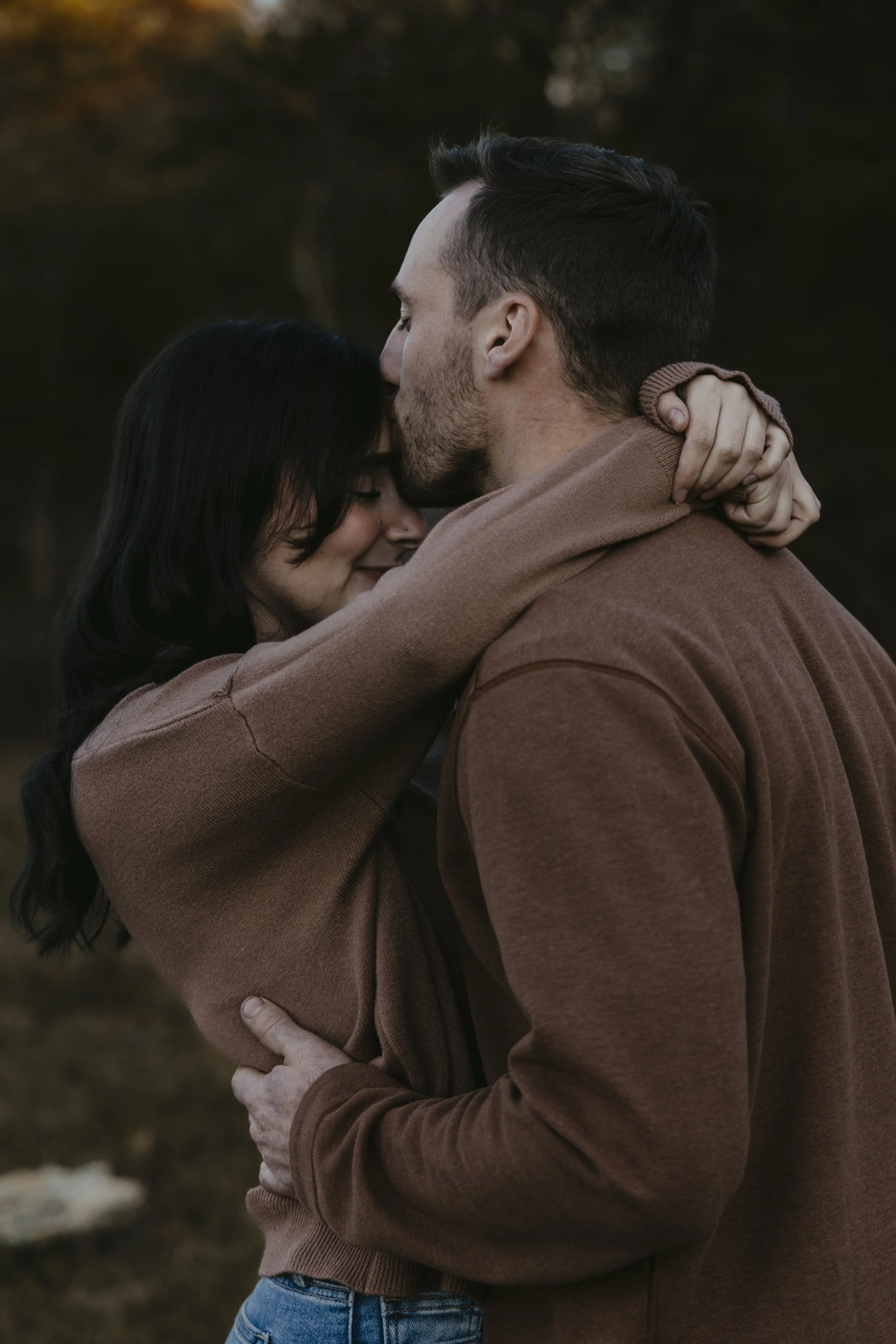 A couple embraces outdoors, with the man kissing the woman's forehead, both with eyes closed and smiling gently.