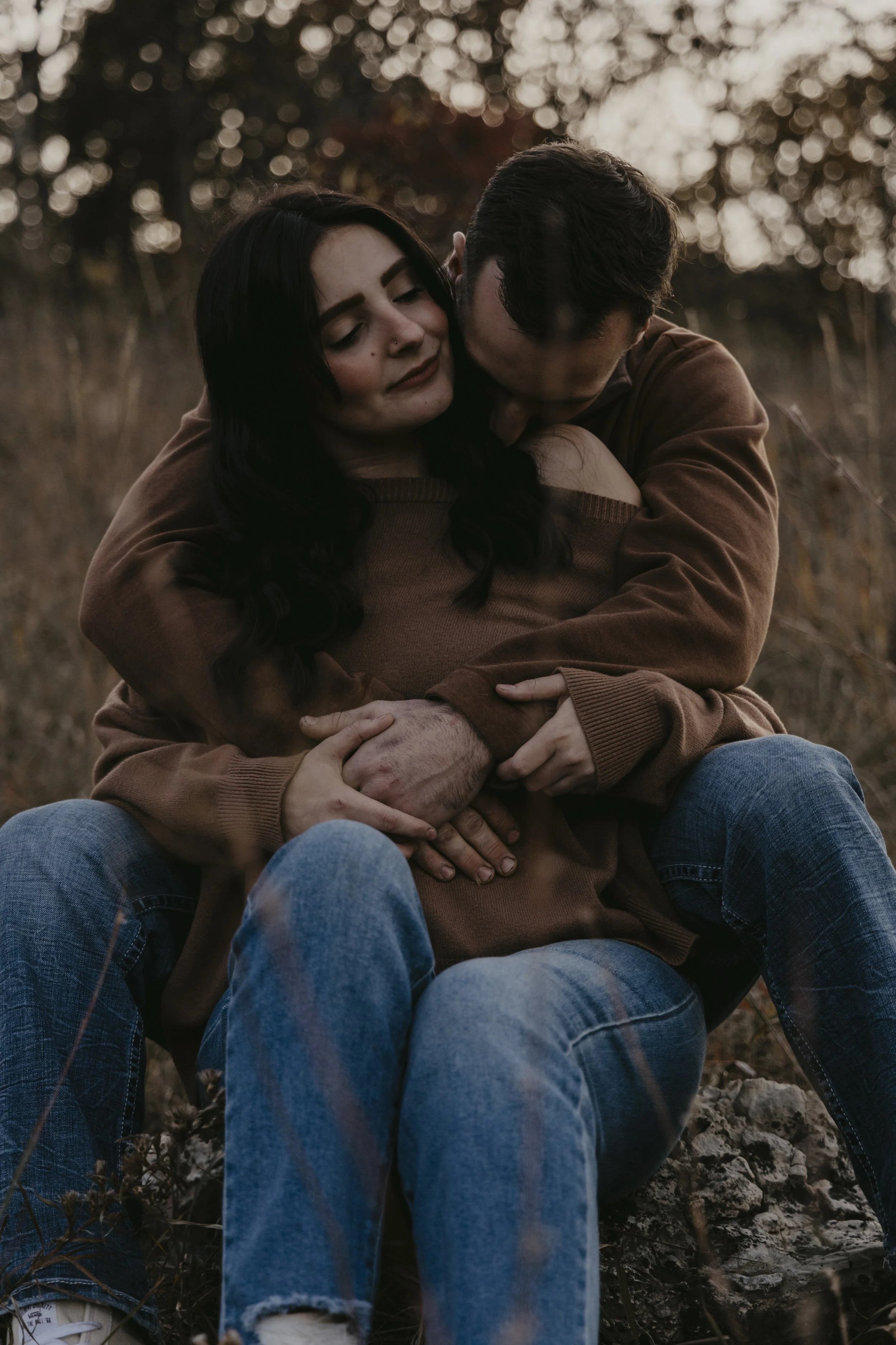 A couple sitting outdoors at sunset, embracing each other, with the woman smiling gently. Both are wearing brown tops and jeans, and the woman has long dark hair. The background features blurred trees and warm lighting.