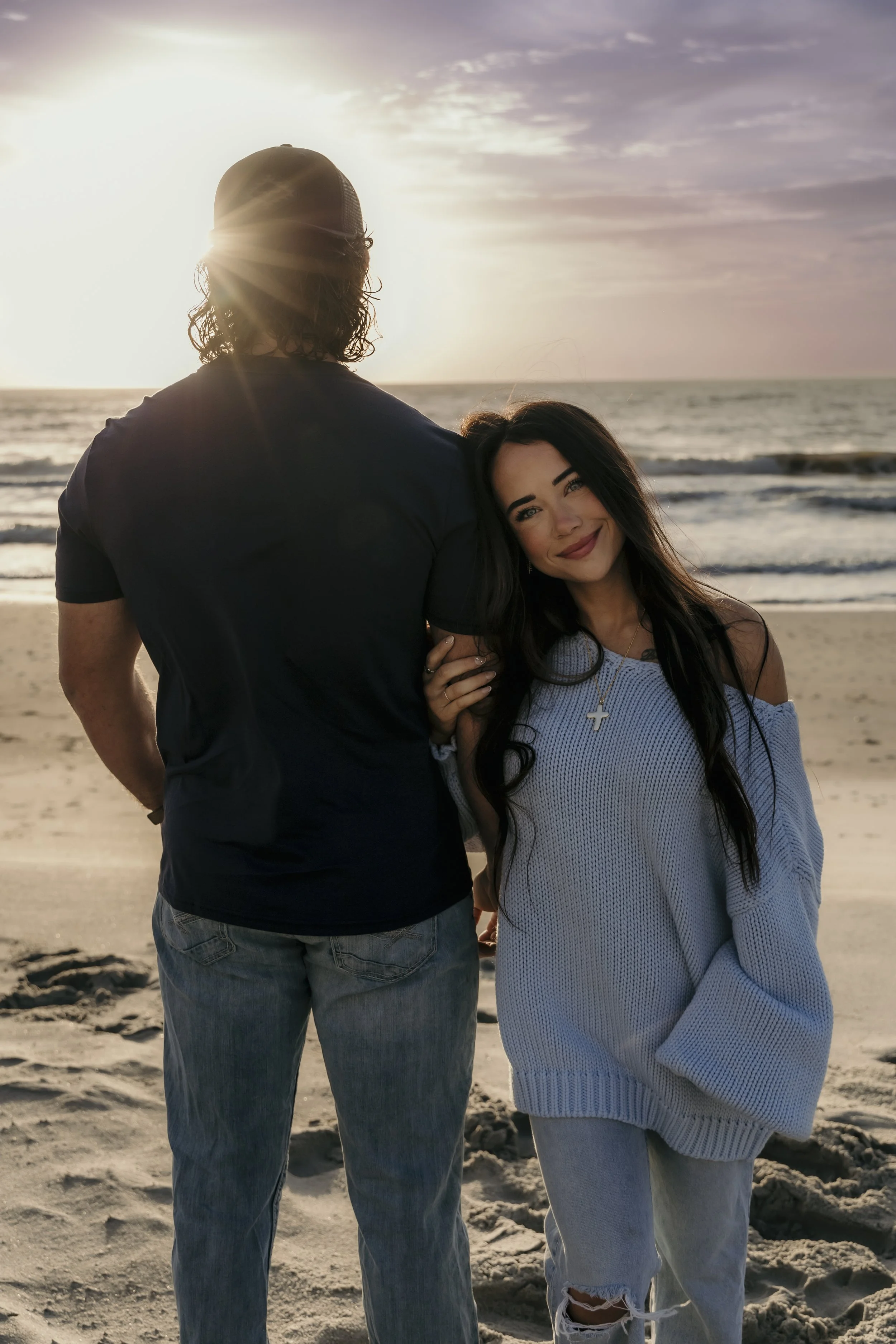 A couple at the beach during sunset, with the woman smiling and leaning her head on the man's shoulder, while the man faces the ocean with the sun setting behind him.