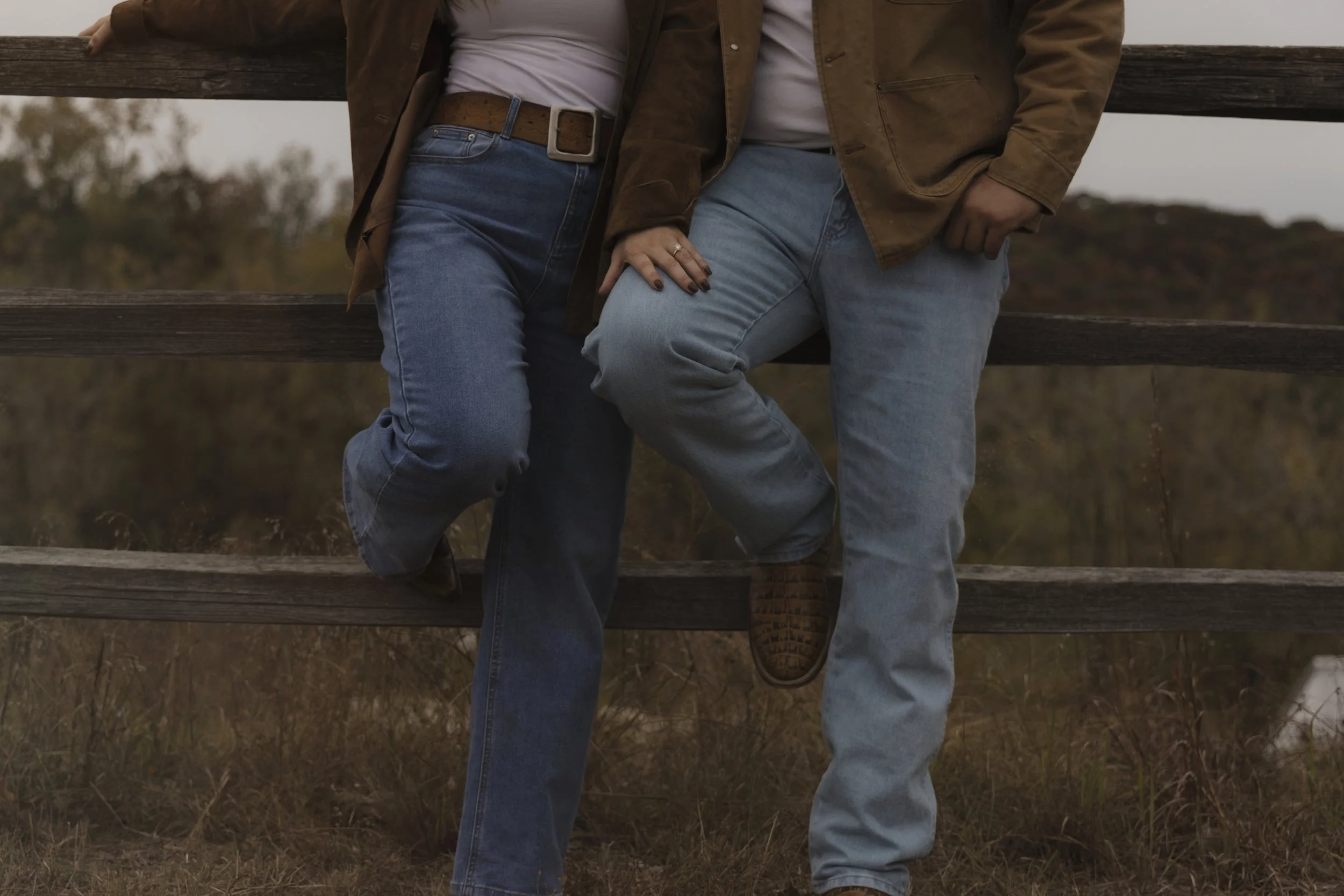 Two people sitting on a wooden fence, with their legs overlapping, outdoors on a cloudy day.