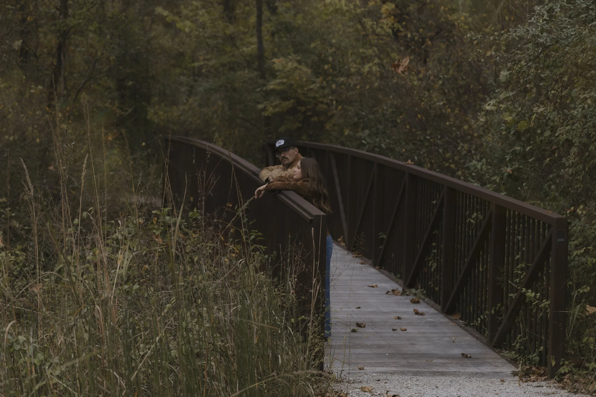 A man and a young girl are leaning on the railing of a wooden bridge in a lush, autumn-colored forest.