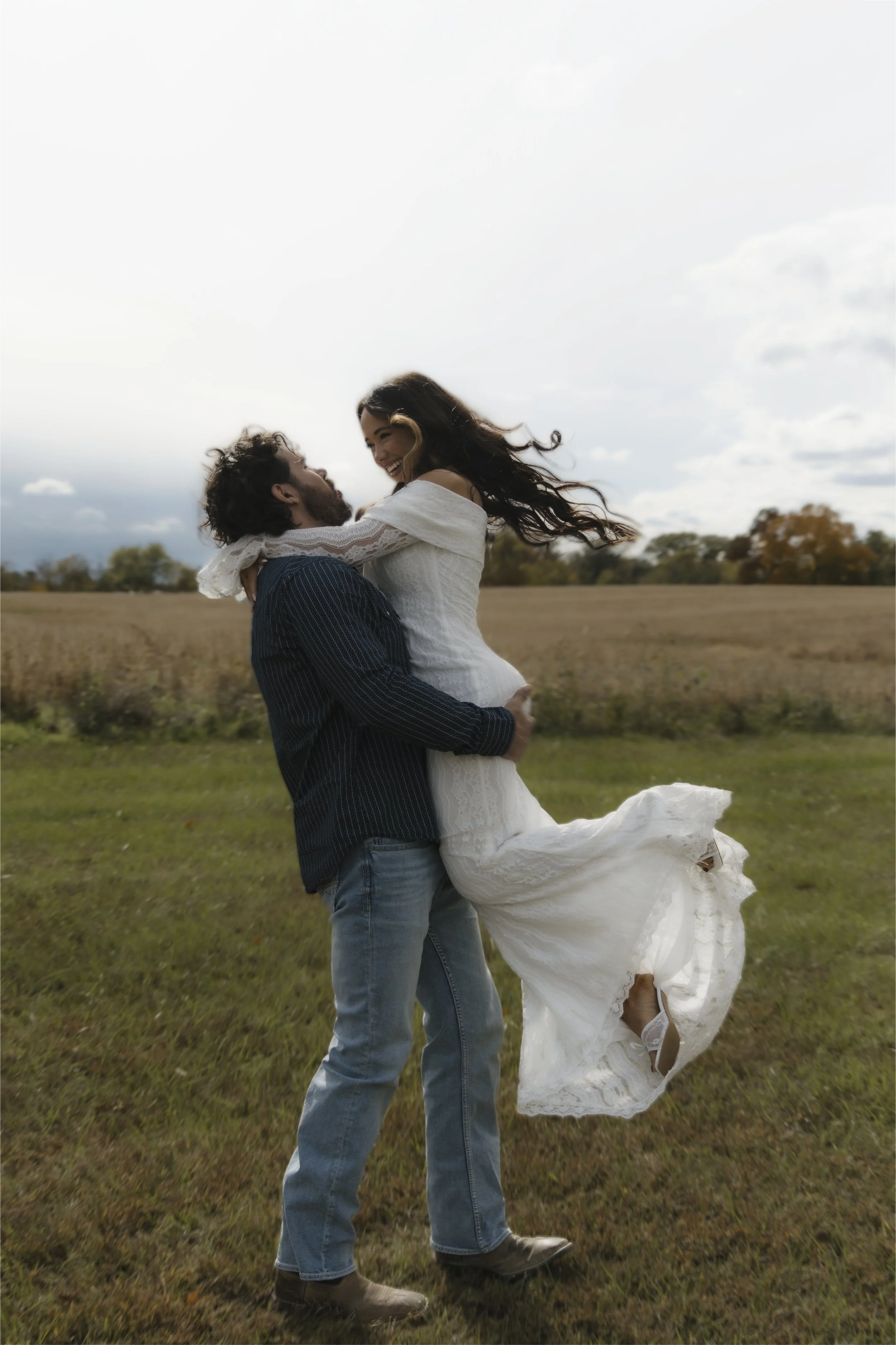 A man lifting a woman in a white dress in a grassy field during daytime, with cloudy sky and trees in the background.
