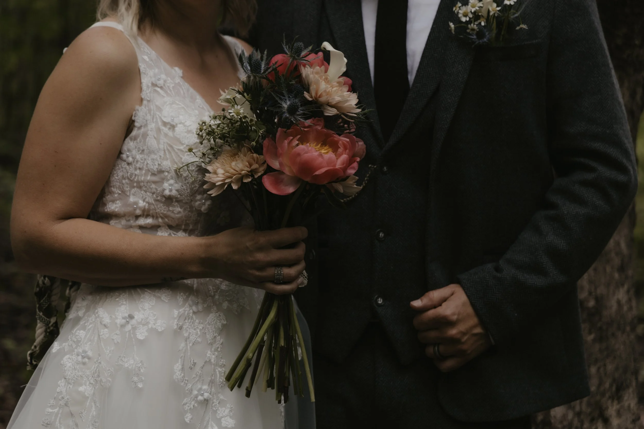 A bride holding a bouquet of pink, cream, and dark blue flowers standing next to a groom in a dark suit outdoors.