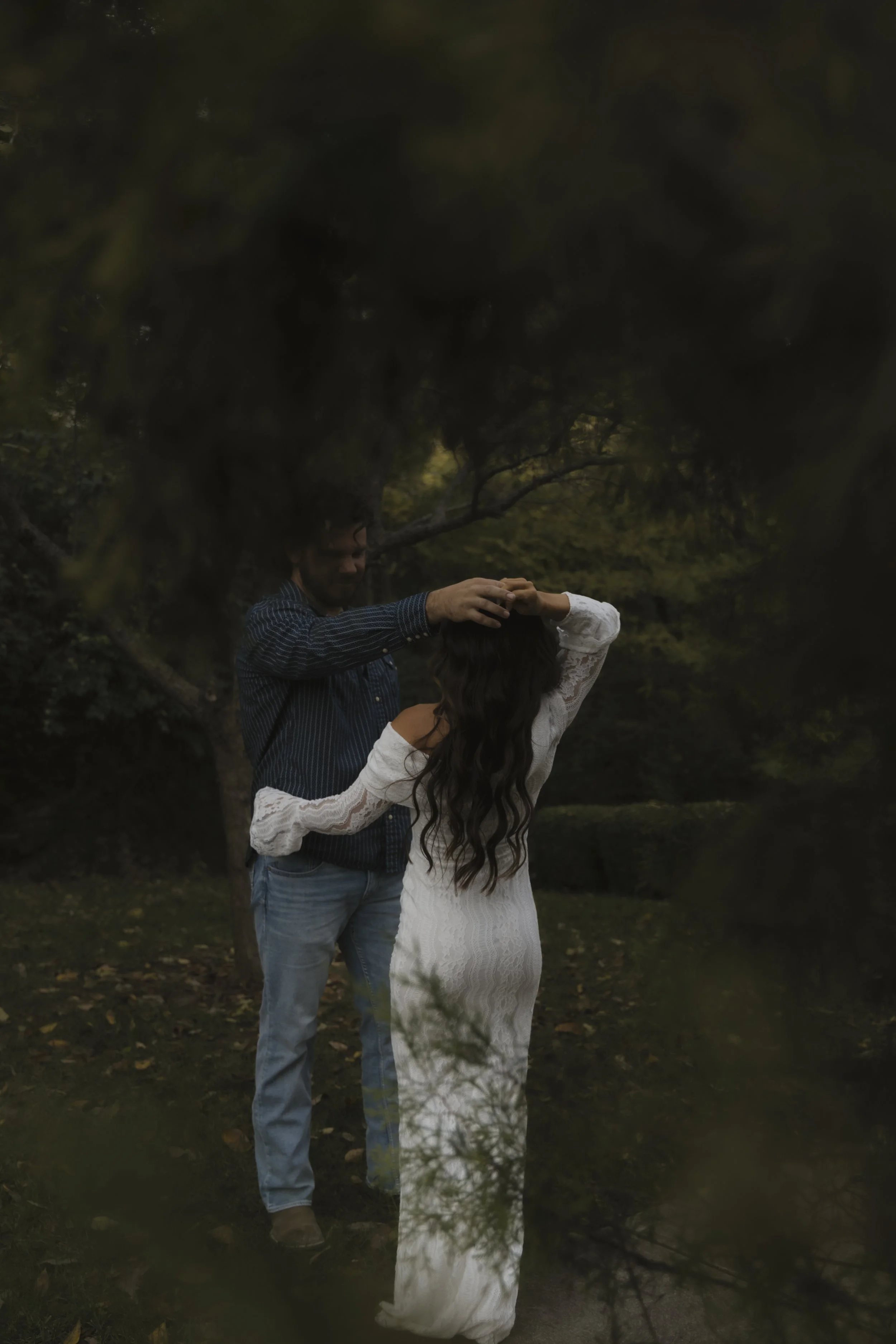 A man and woman standing outdoors in a wooded area, the man touching the woman's head as she faces away from him and adjusts her hair, both dressed in casual clothing.