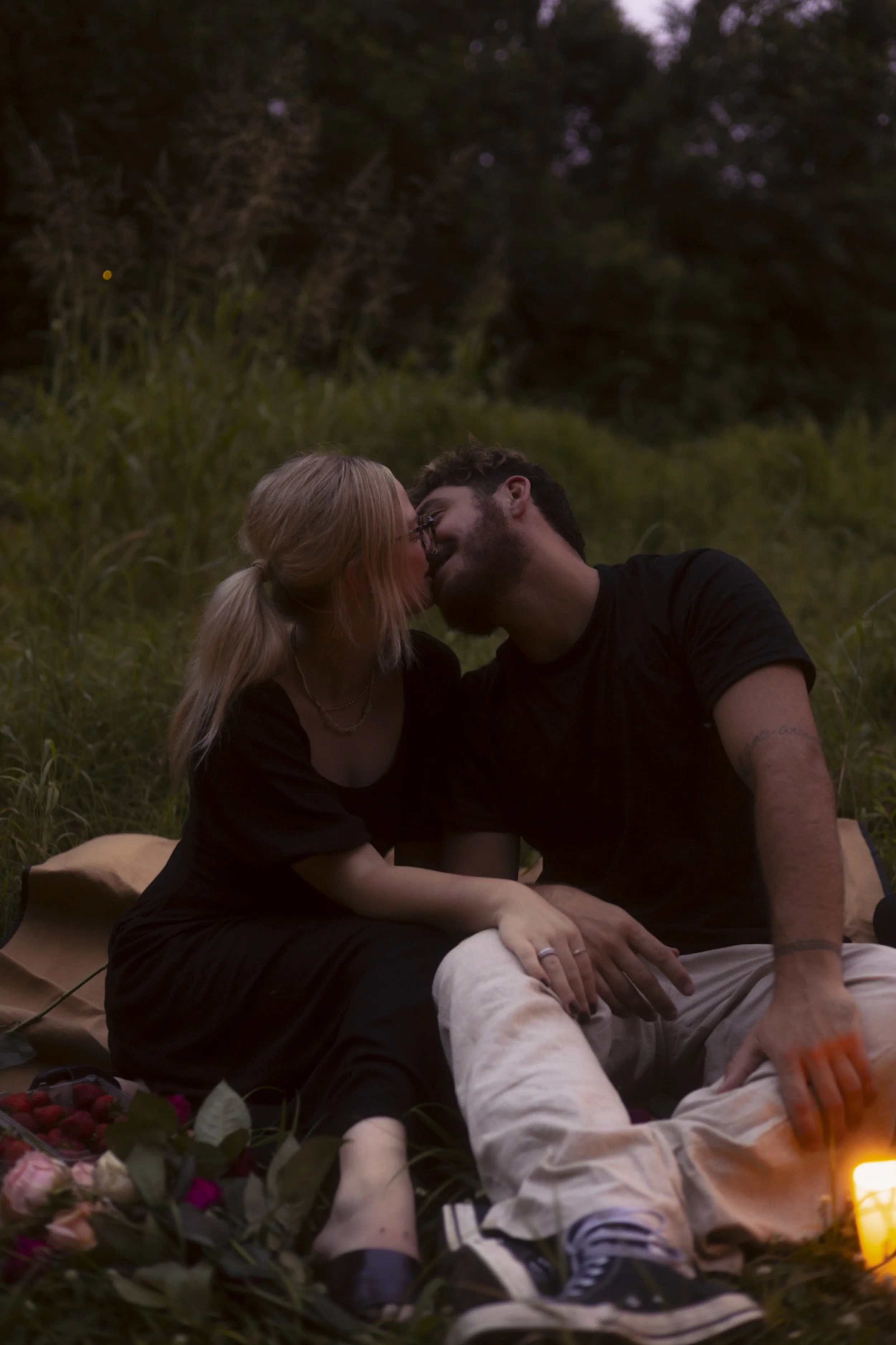 A couple sitting on a blanket in a grassy outdoor area at dusk, leaning in to kiss, with flowers and a lantern nearby.