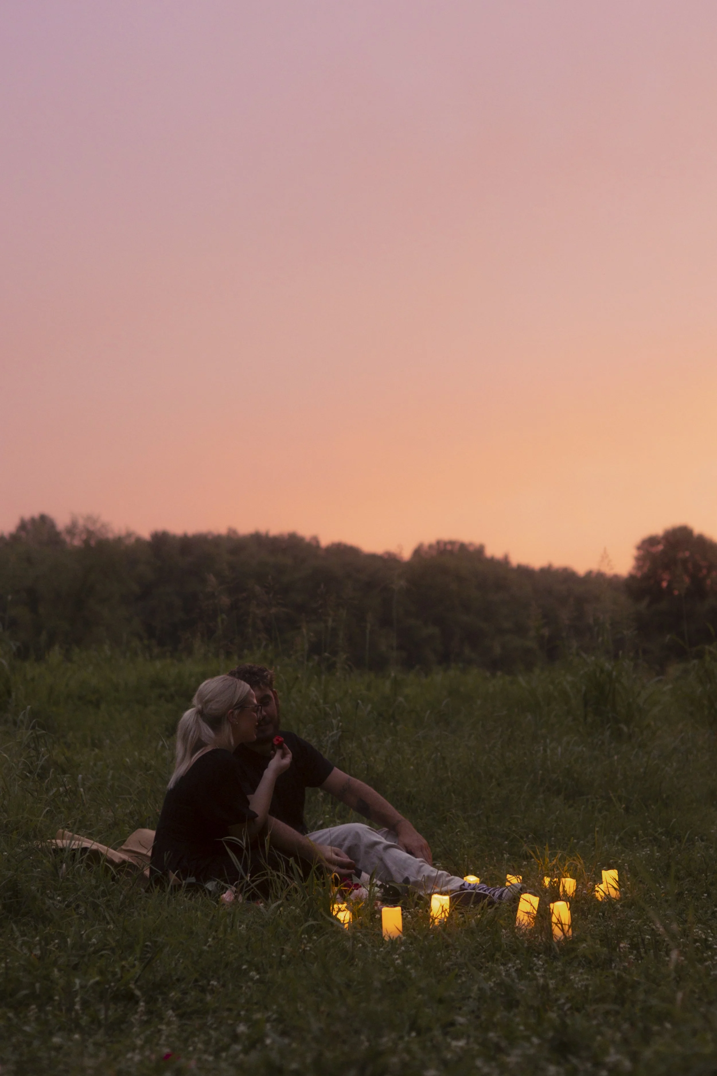A couple sitting on grass at sunset, surrounded by glowing candles, with the woman holding a rose.
