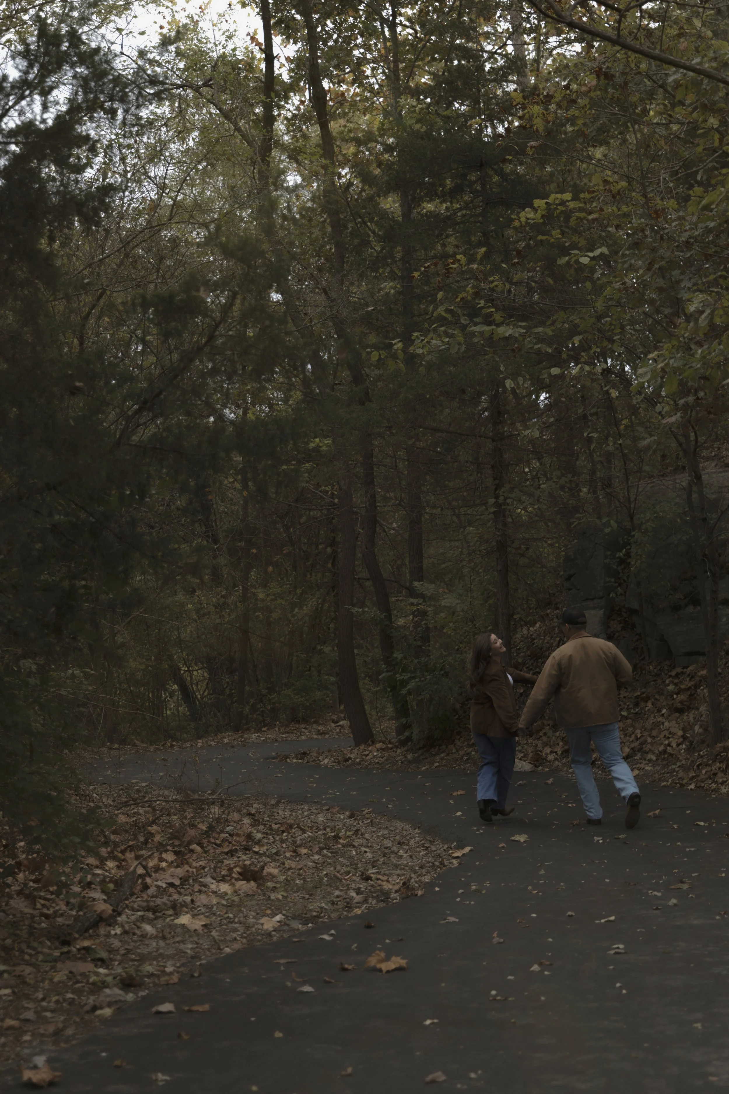 A couple holding hands while walking on a winding forest trail surrounded by trees with fallen leaves.