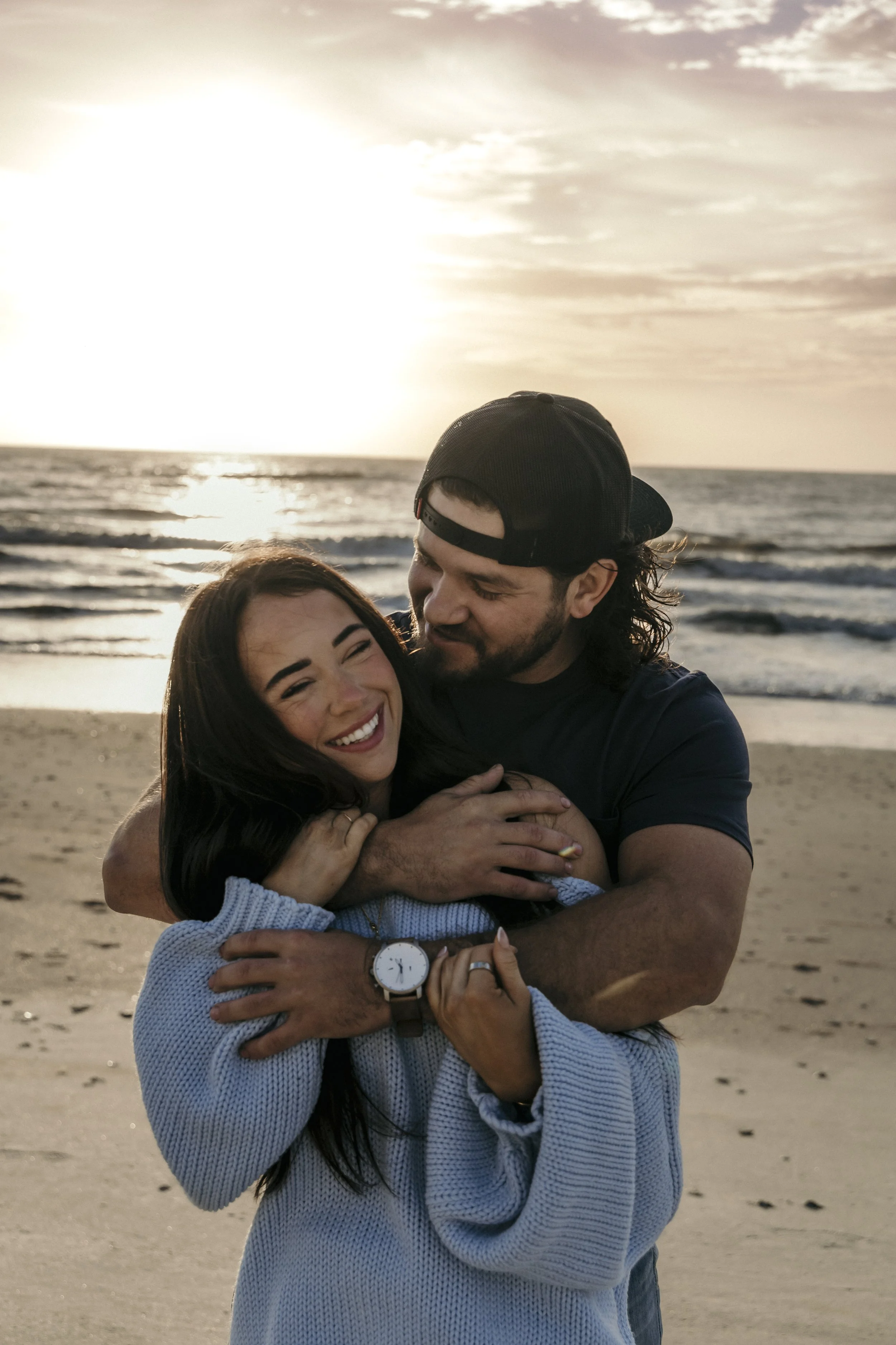 A couple hugging on a beach during sunset, smiling and enjoying each other's company.