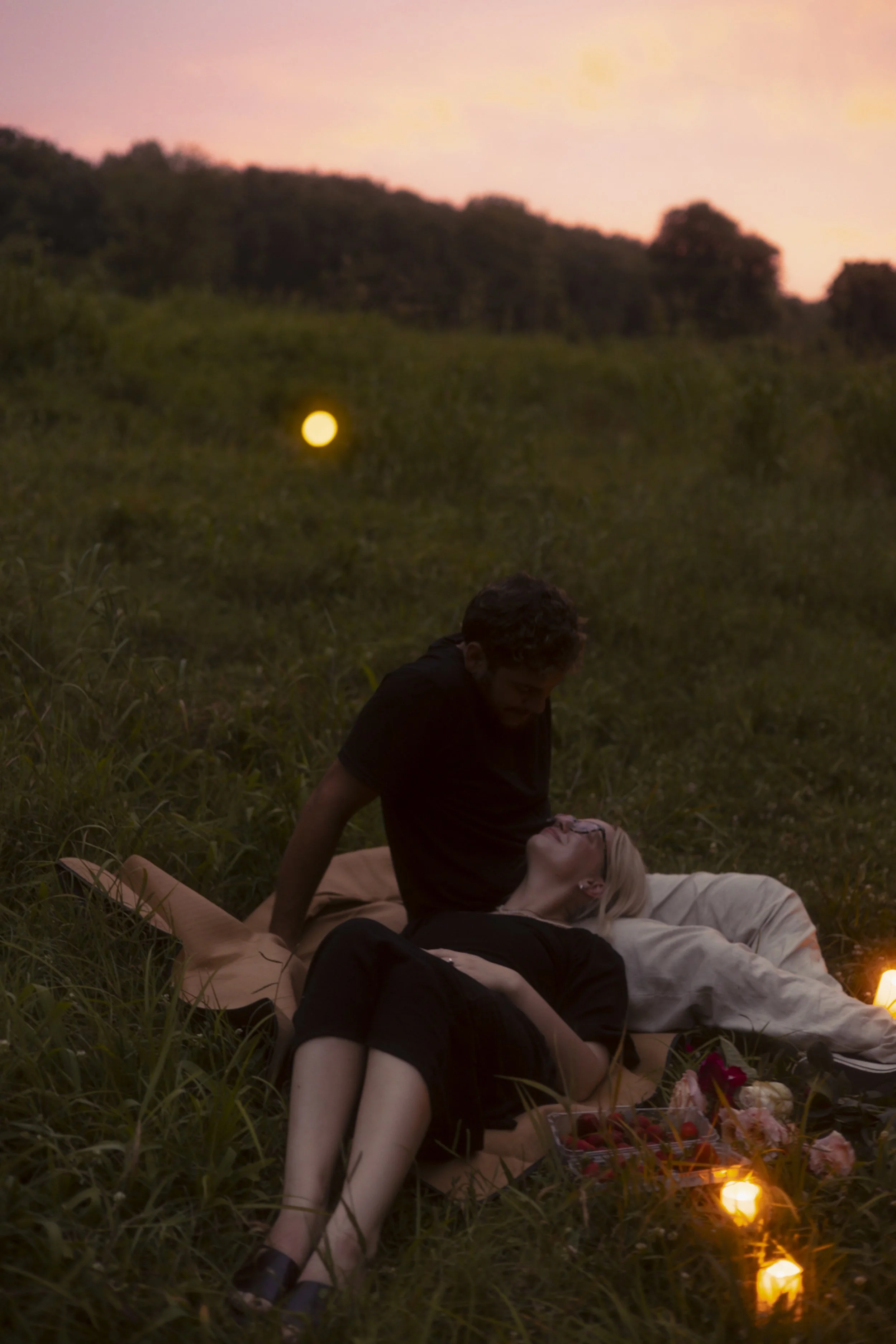 A couple is lying on a blanket in a grassy field during sunset, with candles, flowers, and a platter of strawberries nearby.