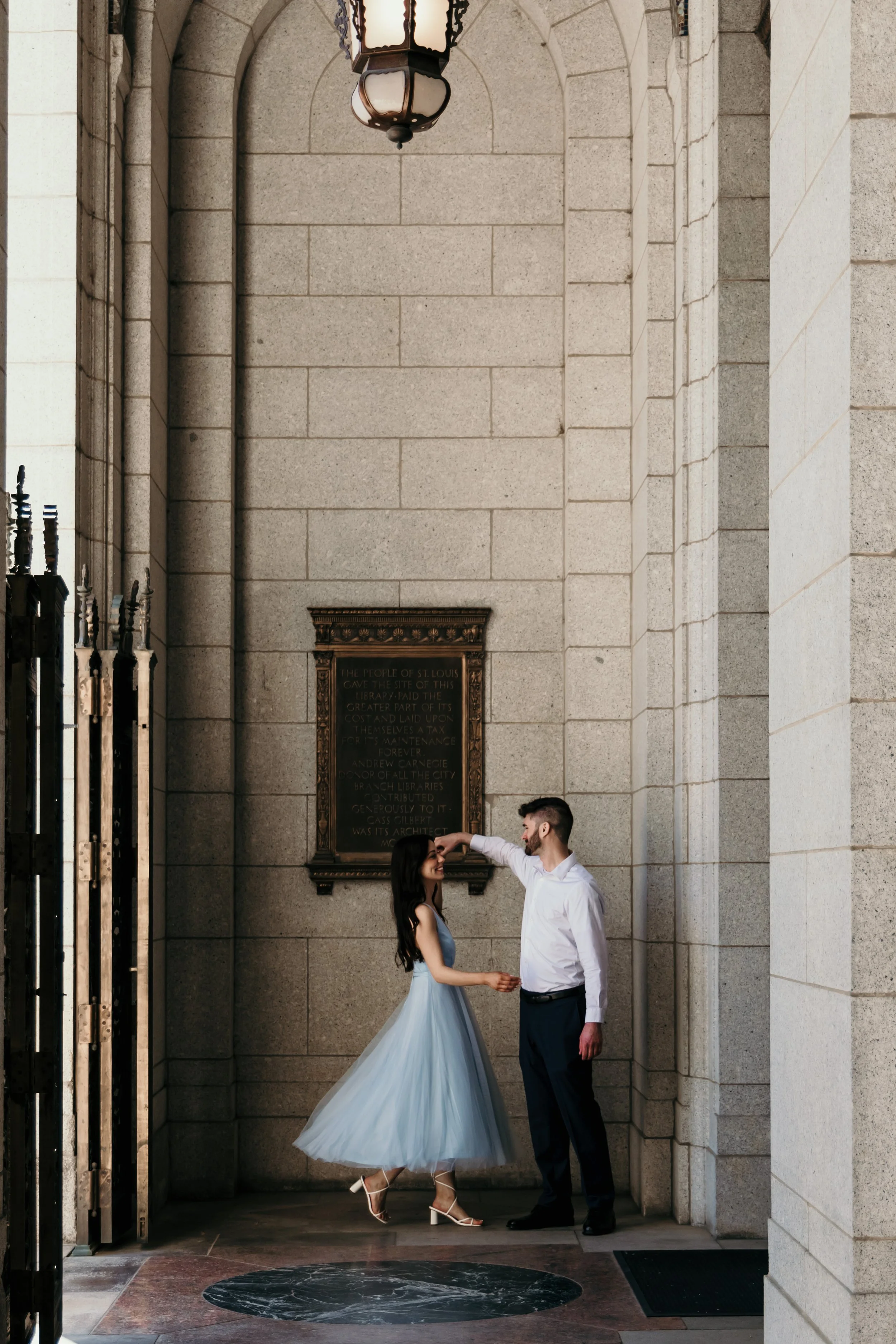 A man and woman dancing together in a stone hallway with a metal gate on one side and a plaque on the wall behind them.