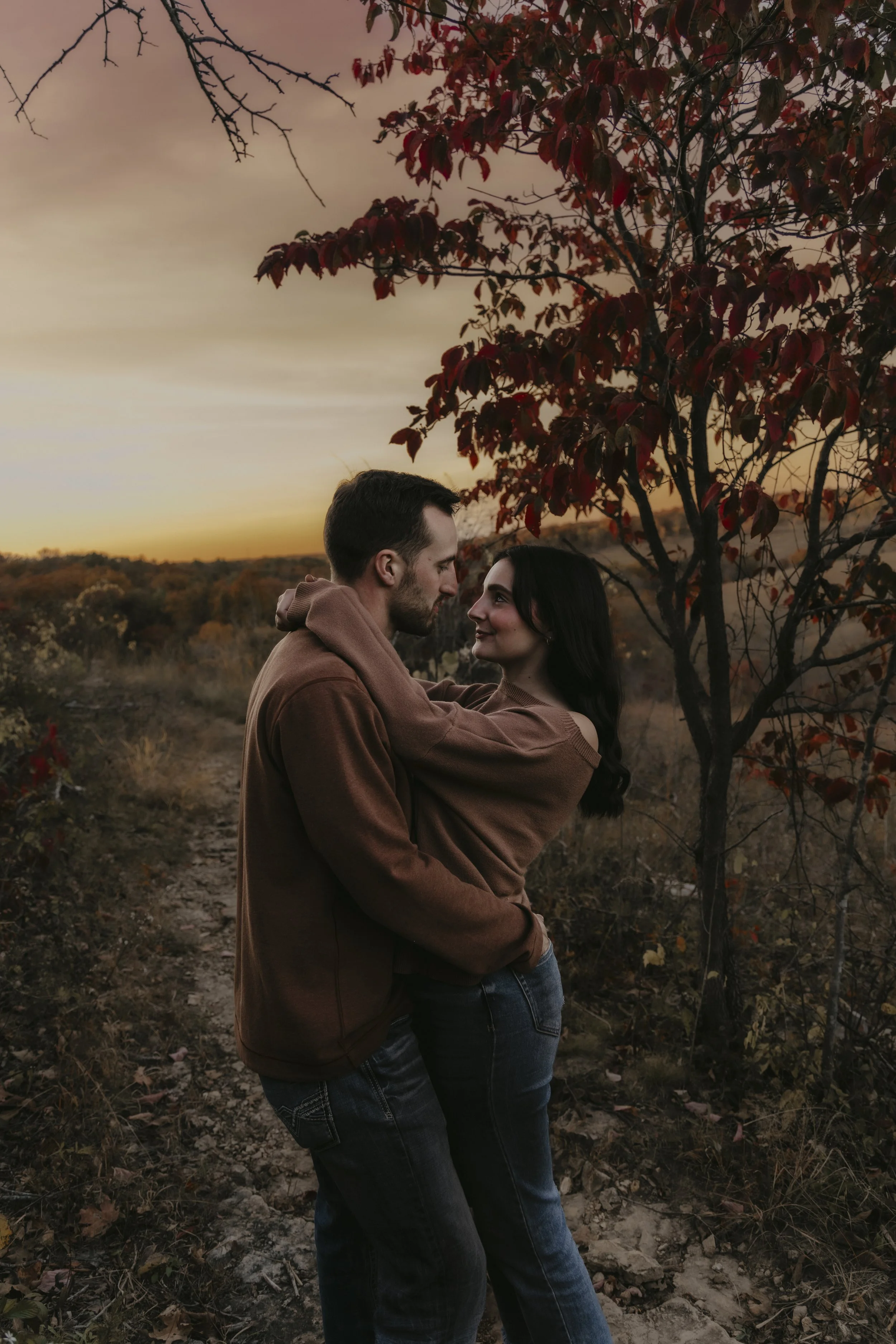 A couple embracing outdoors during sunset, standing on a dirt path near a tree with red leaves in fall.