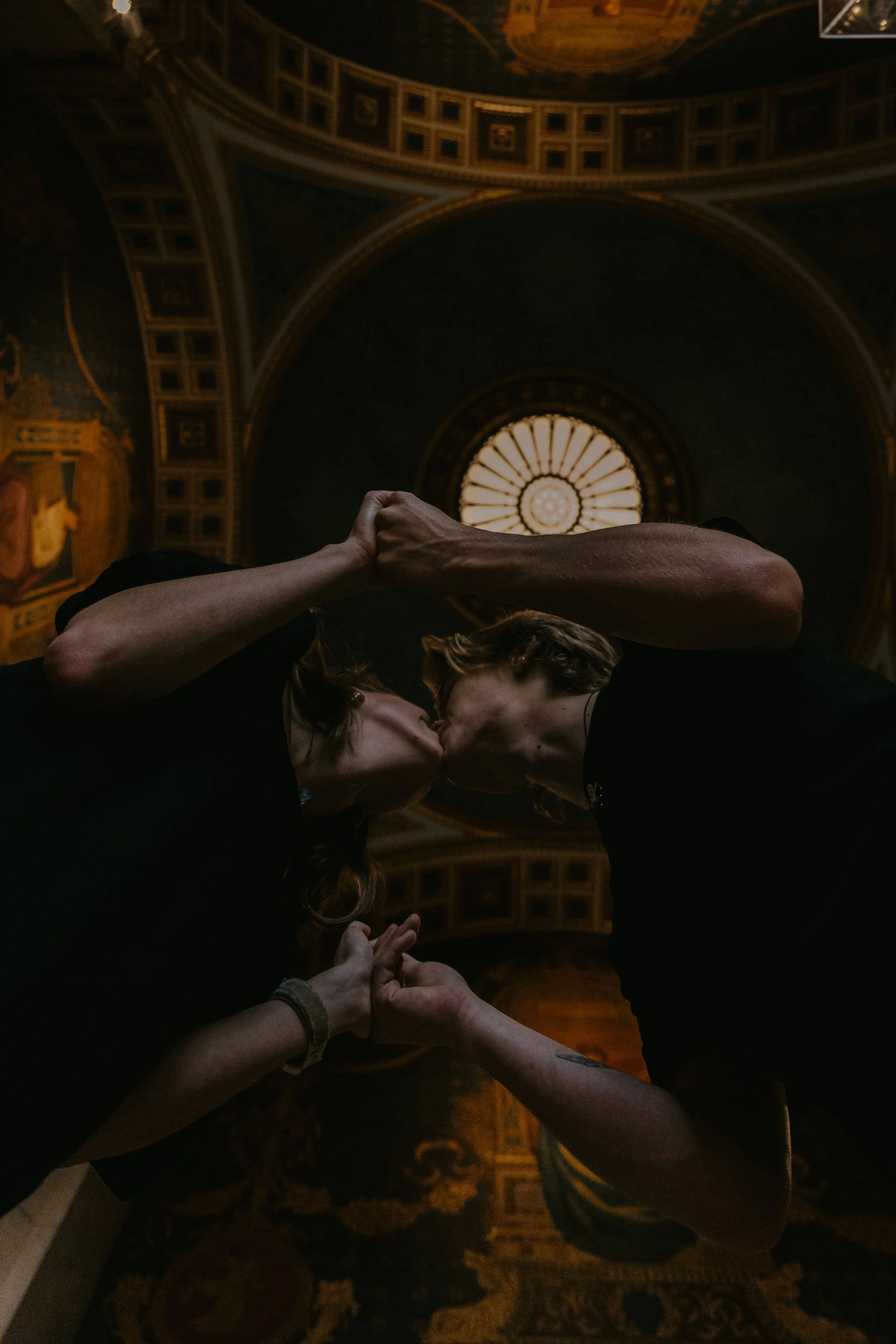 Two people are kissing under a dome ceiling, holding hands and touching foreheads, in a dimly lit ornate room.