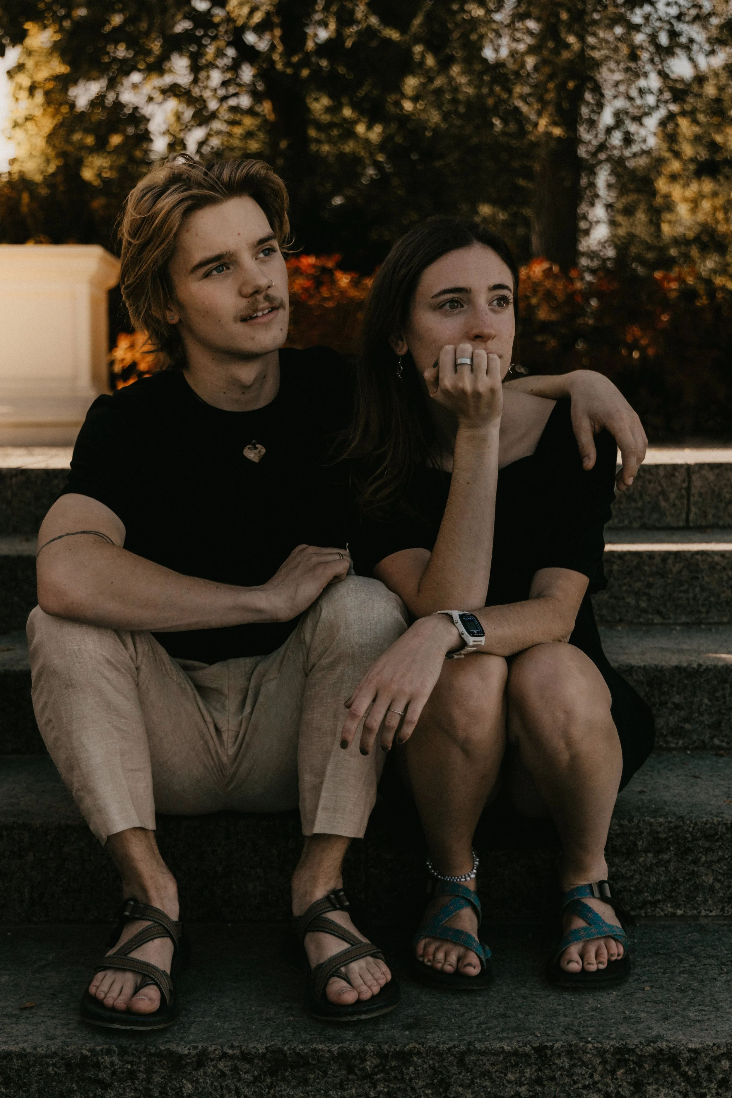 A young man and woman sitting on steps outdoors during sunset, with the man having his arm around the woman's shoulder, both looking thoughtful.