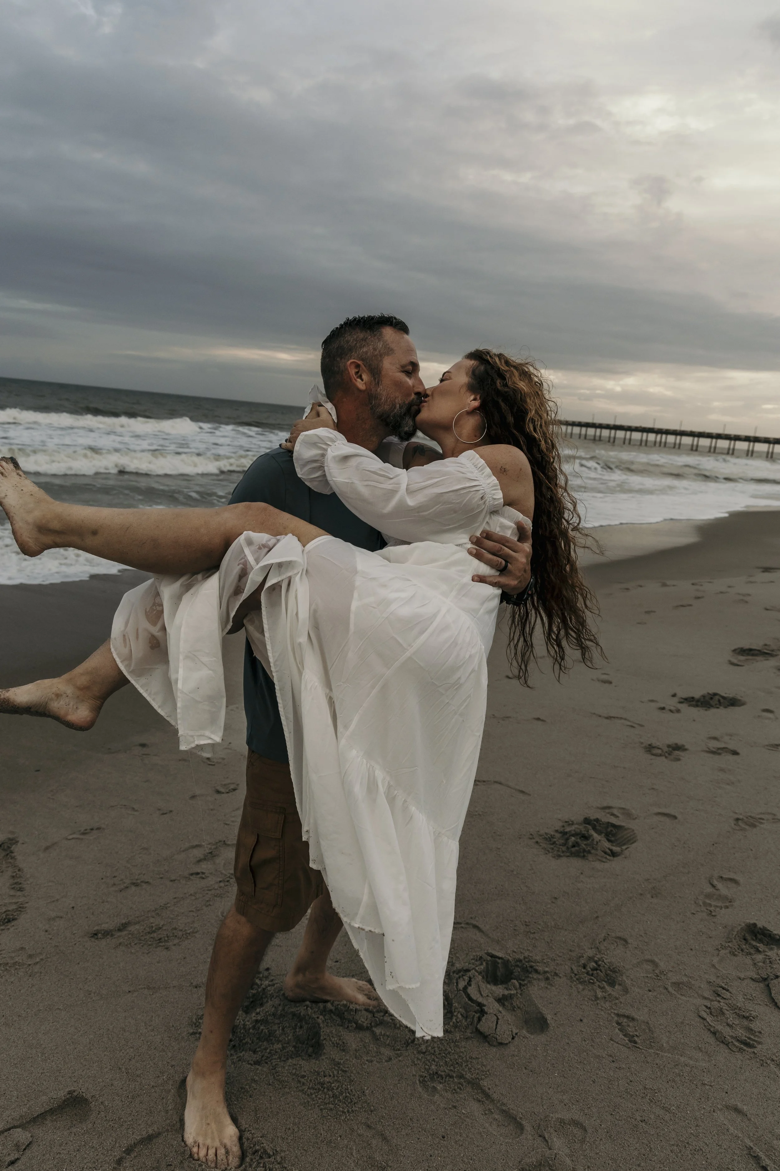 A man carrying a woman in a white dress on the beach, both kissing, with a cloudy sky and a pier in the background.