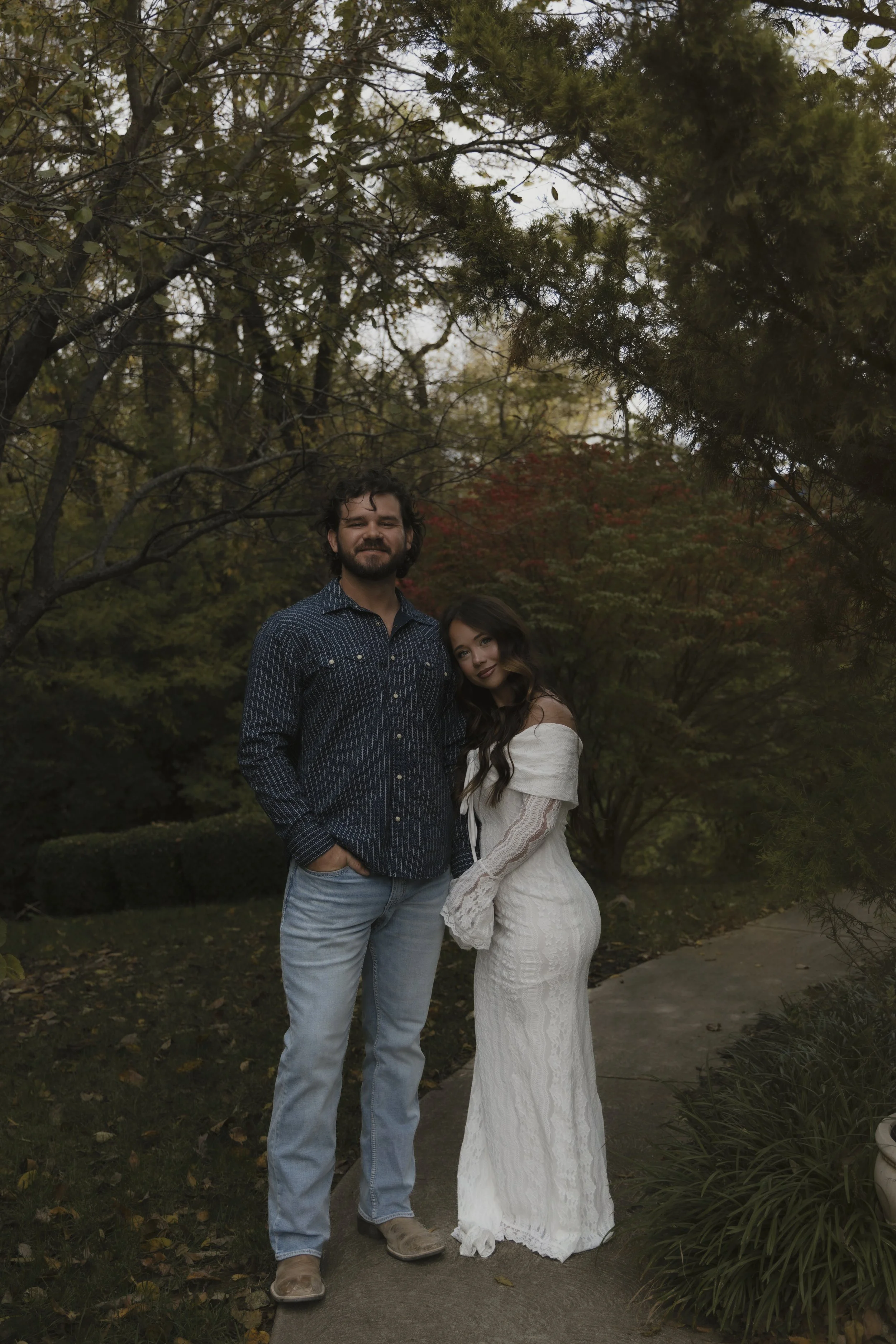 A man and woman standing outdoors on a sidewalk in front of trees with autumn foliage, smiling at the camera.