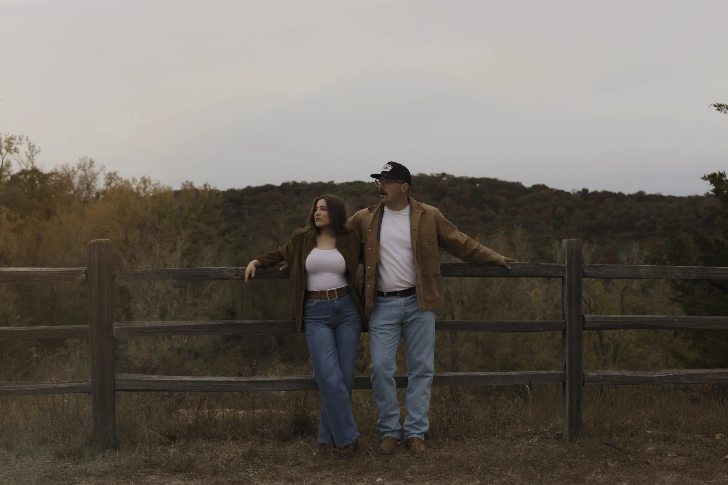 A man and woman standing and leaning on a wooden fence outdoors, with trees and hills in the background during late afternoon or early evening.