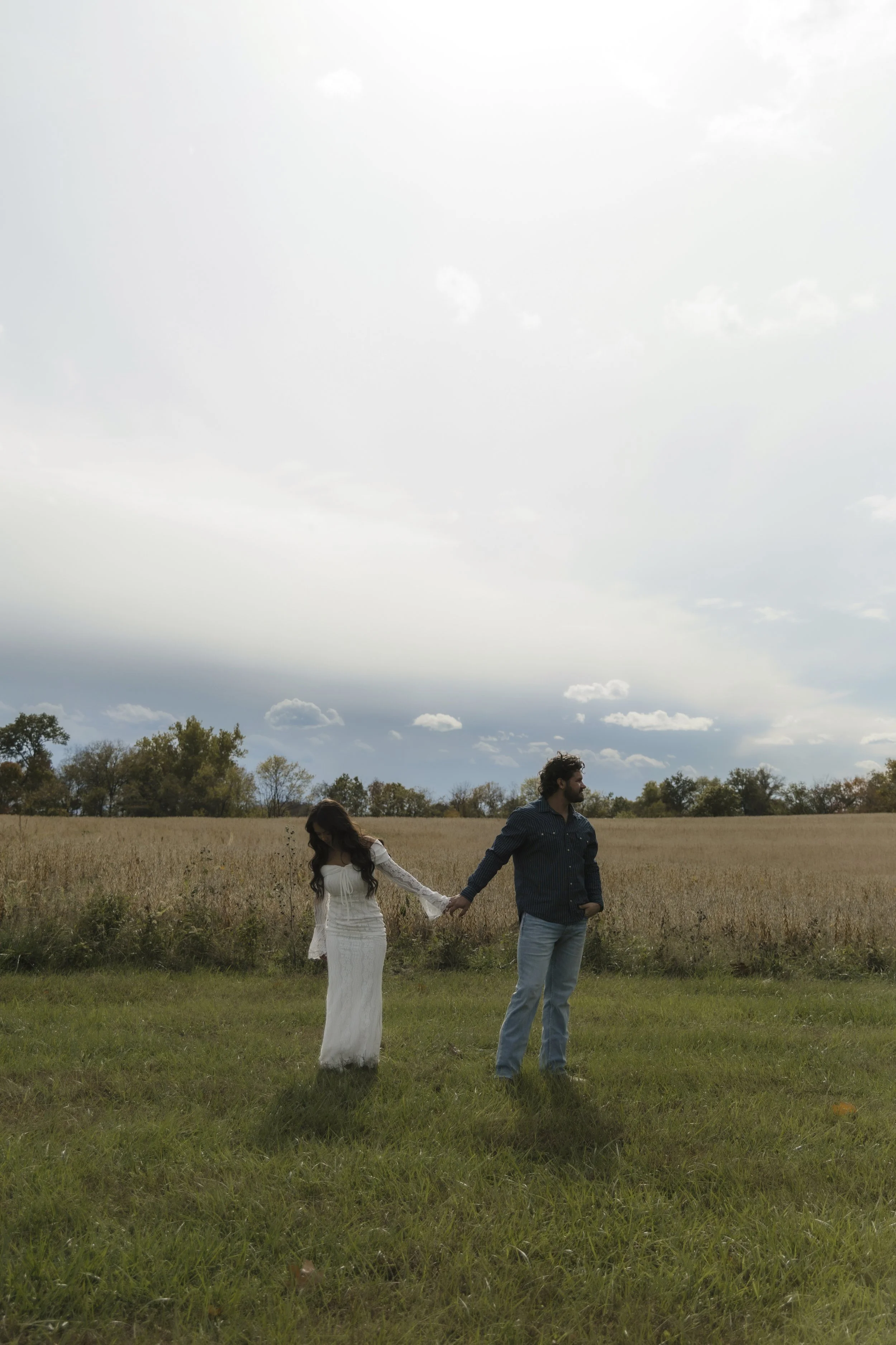 A couple holding hands in a field with a cloudy sky overhead.
