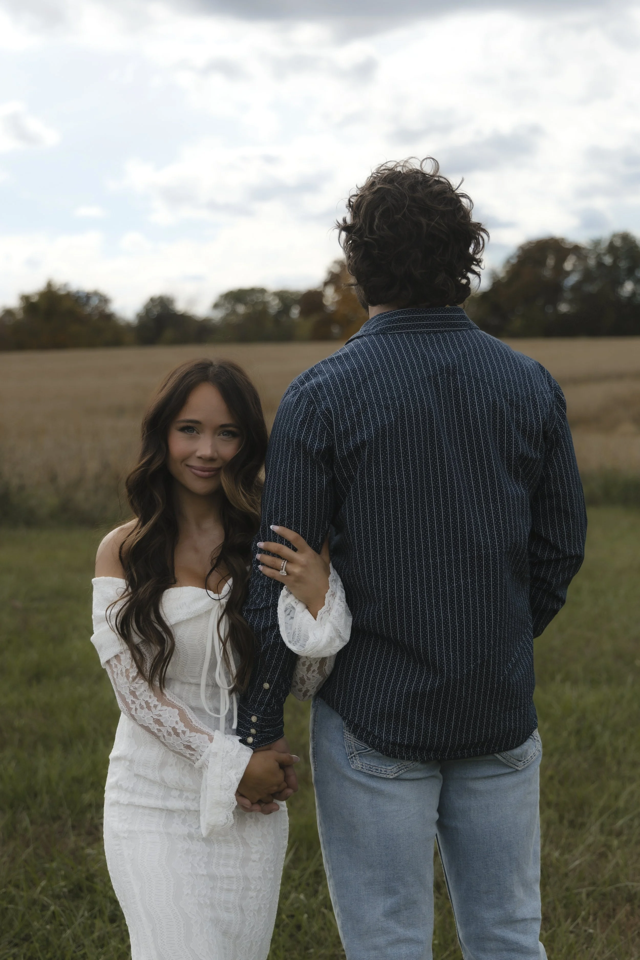 A woman in a white dress holding hands with a man in jeans and a blue striped shirt in an outdoor field.