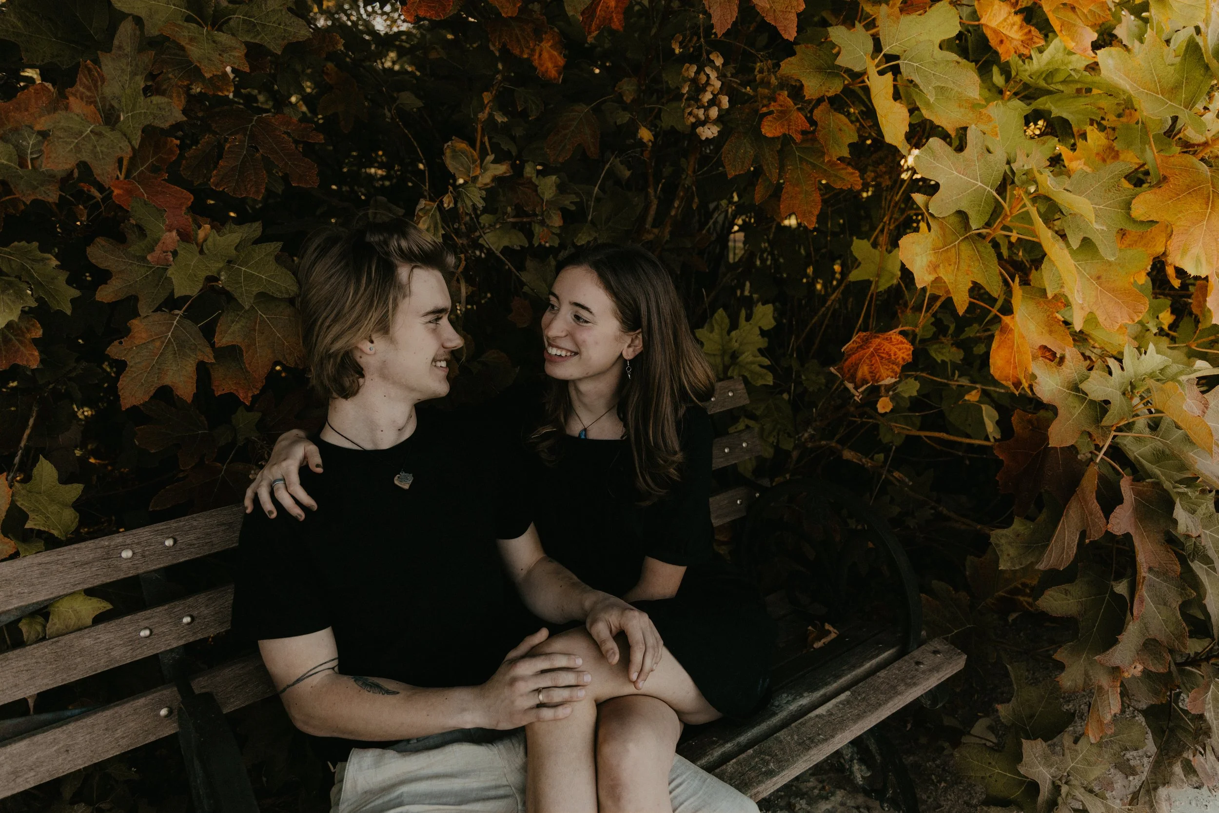 A young couple sitting on a wooden bench surrounded by autumn leaves, sharing a tender moment and smiling at each other.
