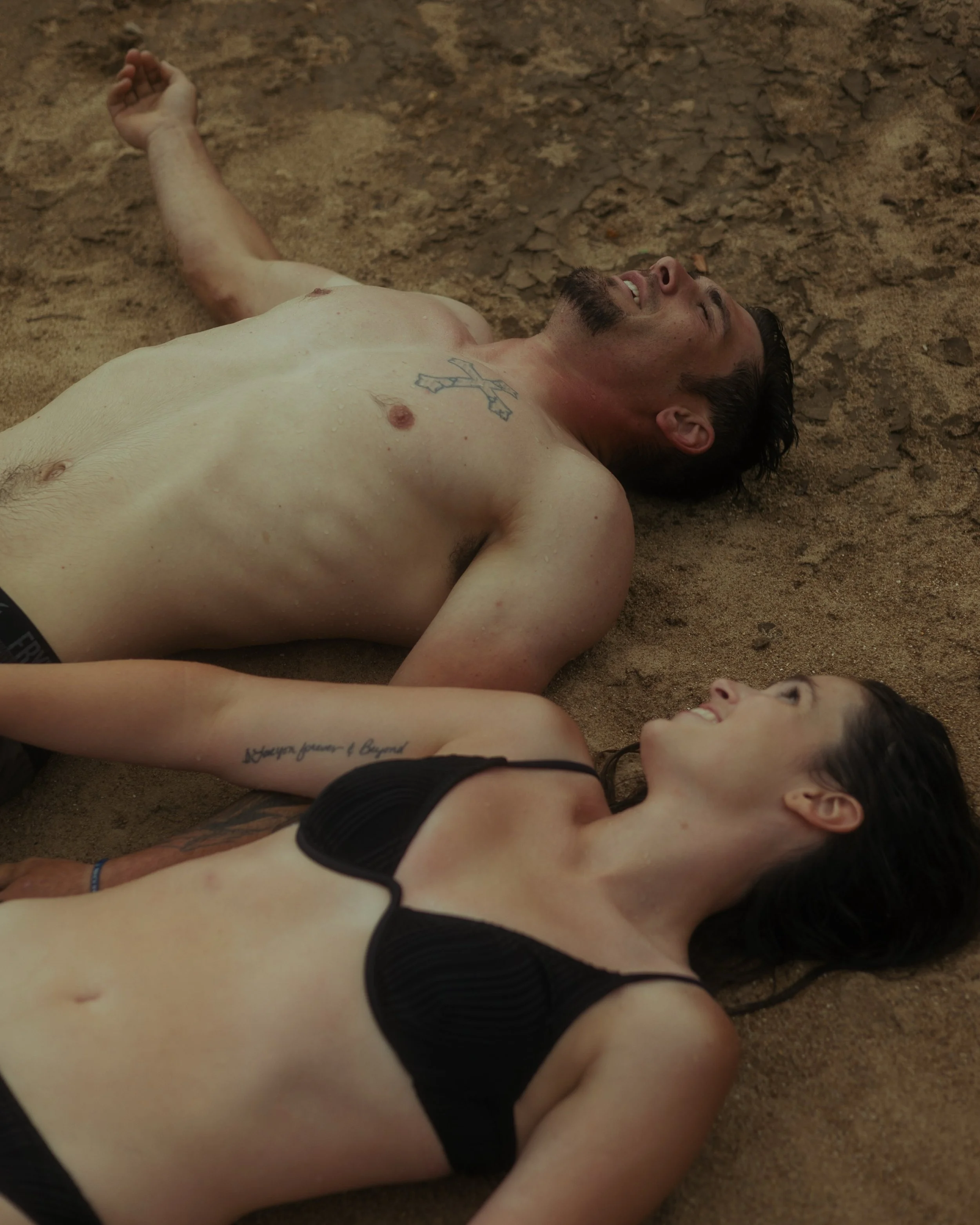 A shirtless man and a woman in a black bikini lying on sandy ground, looking at each other and smiling.