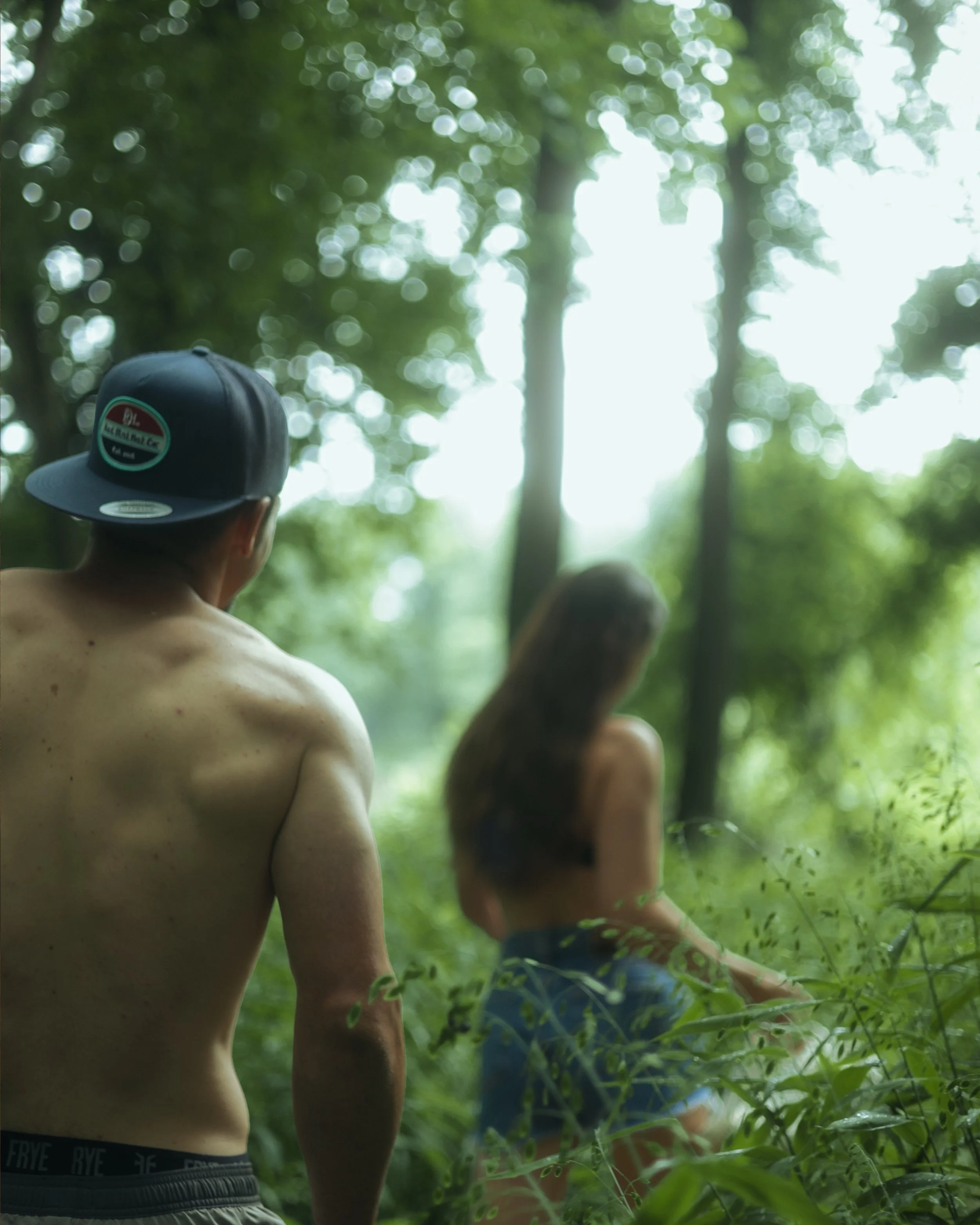 Shirtless man with a backwards cap standing in a green, wooded area with a woman sitting on the ground in front of him, also shirtless, with long hair and jeans, surrounded by dense foliage and tall trees.