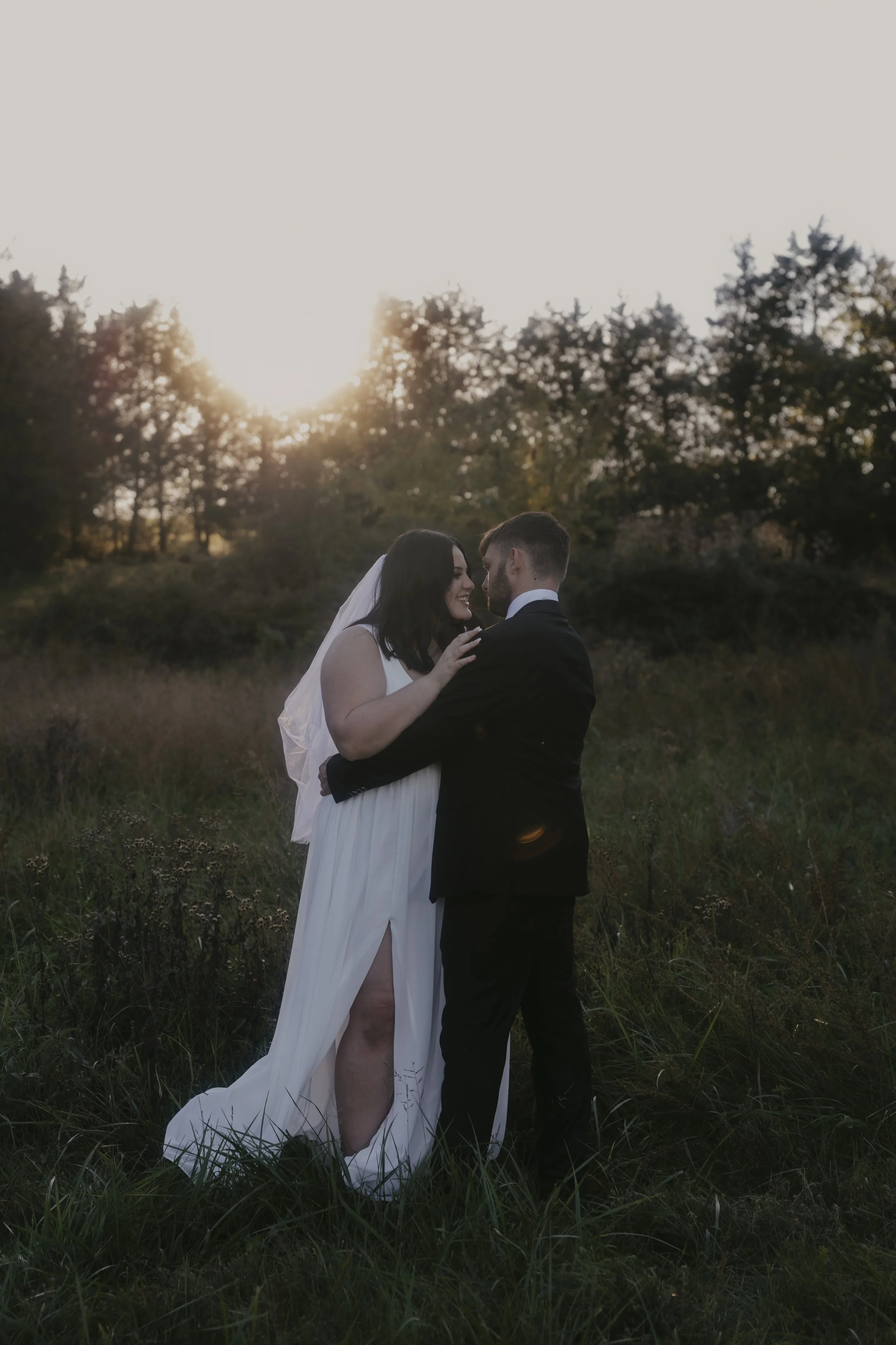 A bride and groom in wedding attire embrace outdoors in a grassy field during sunset.