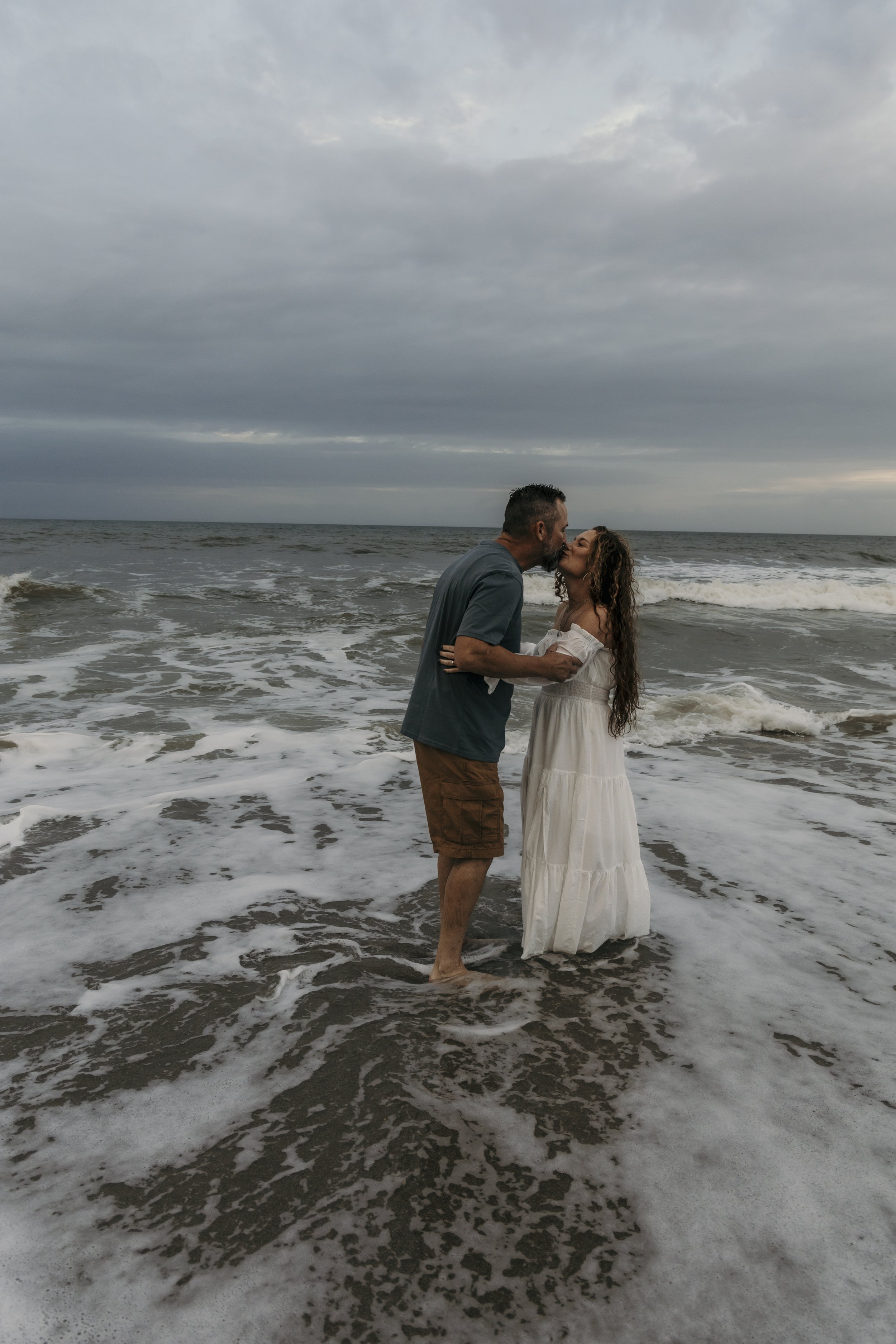 A couple kissing on the beach at sunset, with ocean waves in the background and a cloudy sky overhead.