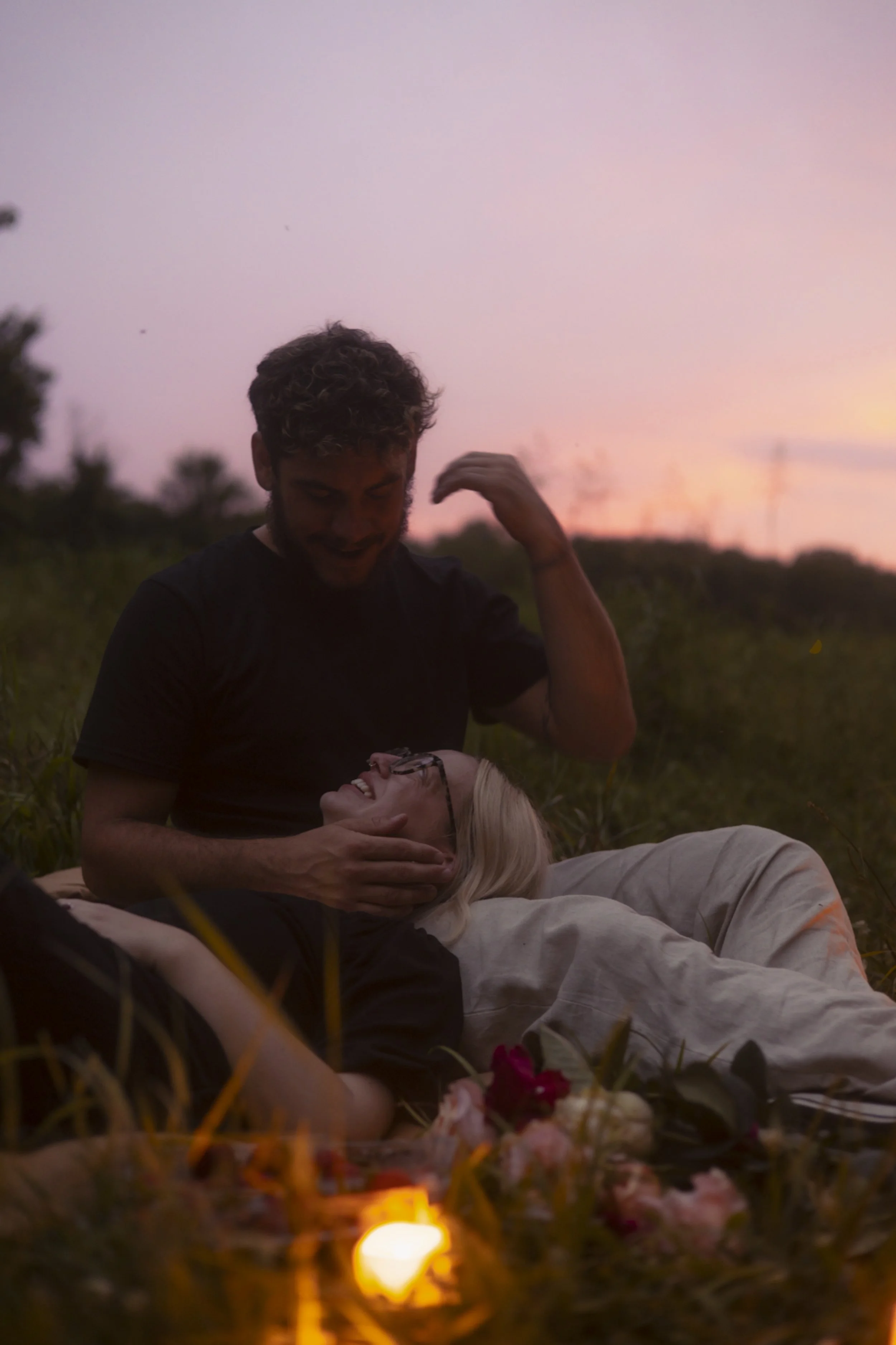 A young man is kneeling on the grass, smiling and gently touching the face of a woman lying on her back among flowers and candles outdoors during sunset.