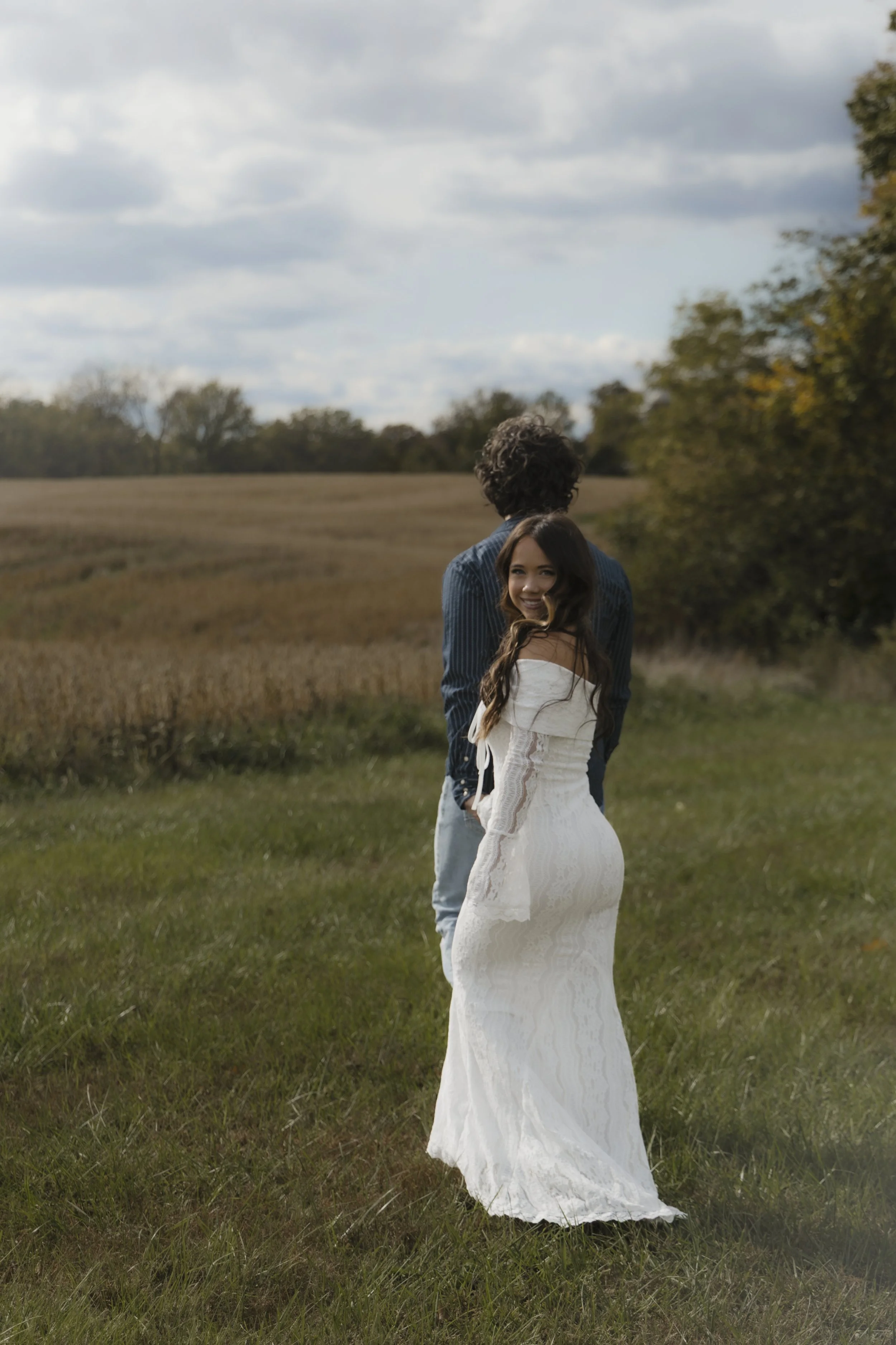 A woman in a white wedding dress smiling and turning back in a grassy field with a man standing behind her, facing away, during daytime sky with clouds.
