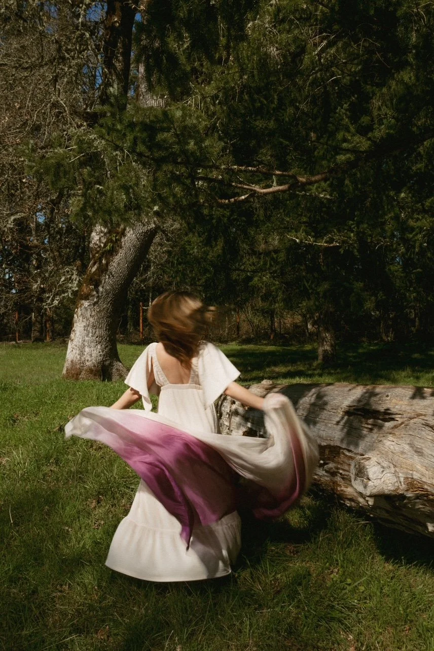 A woman in a white dress spinning outdoors near a fallen log, surrounded by trees and grass. Somatic/embodied movement therapy.