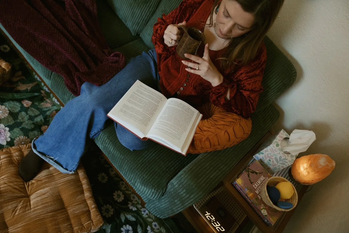 A woman sitting on a green sofa, reading a large open book and drinking from a mug. There is a side table with a tissue box, a decorative salt lamp, a small plant, and some books and magazines. The floor has a floral carpet, and she is wearing jeans 
