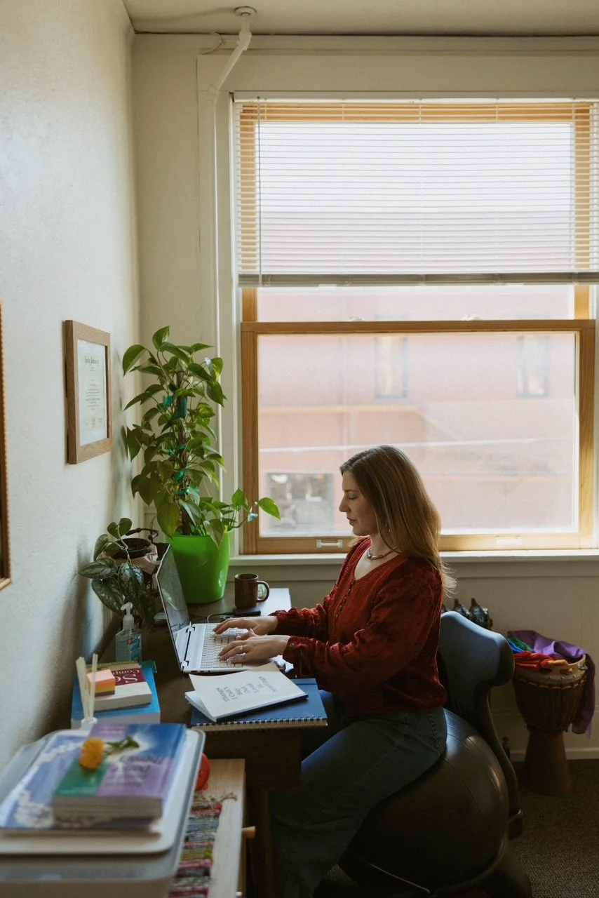 A woman sitting at a desk working on a laptop, with plants, a coffee mug, and papers nearby in a well-lit room with a large window.