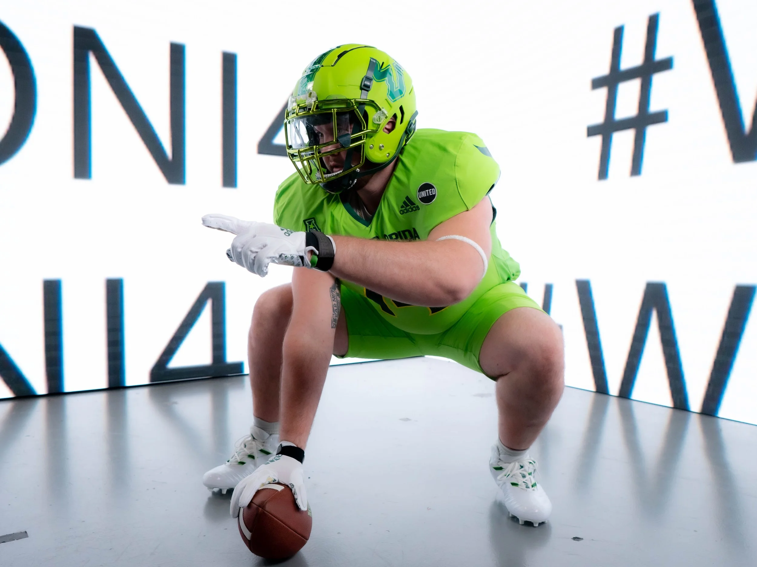 American football player in neon green uniform crouching with one hand on the ground and the other pointing forward, holding a football, wearing a helmet and gloves, on a field with a bright digital scoreboard in the background.