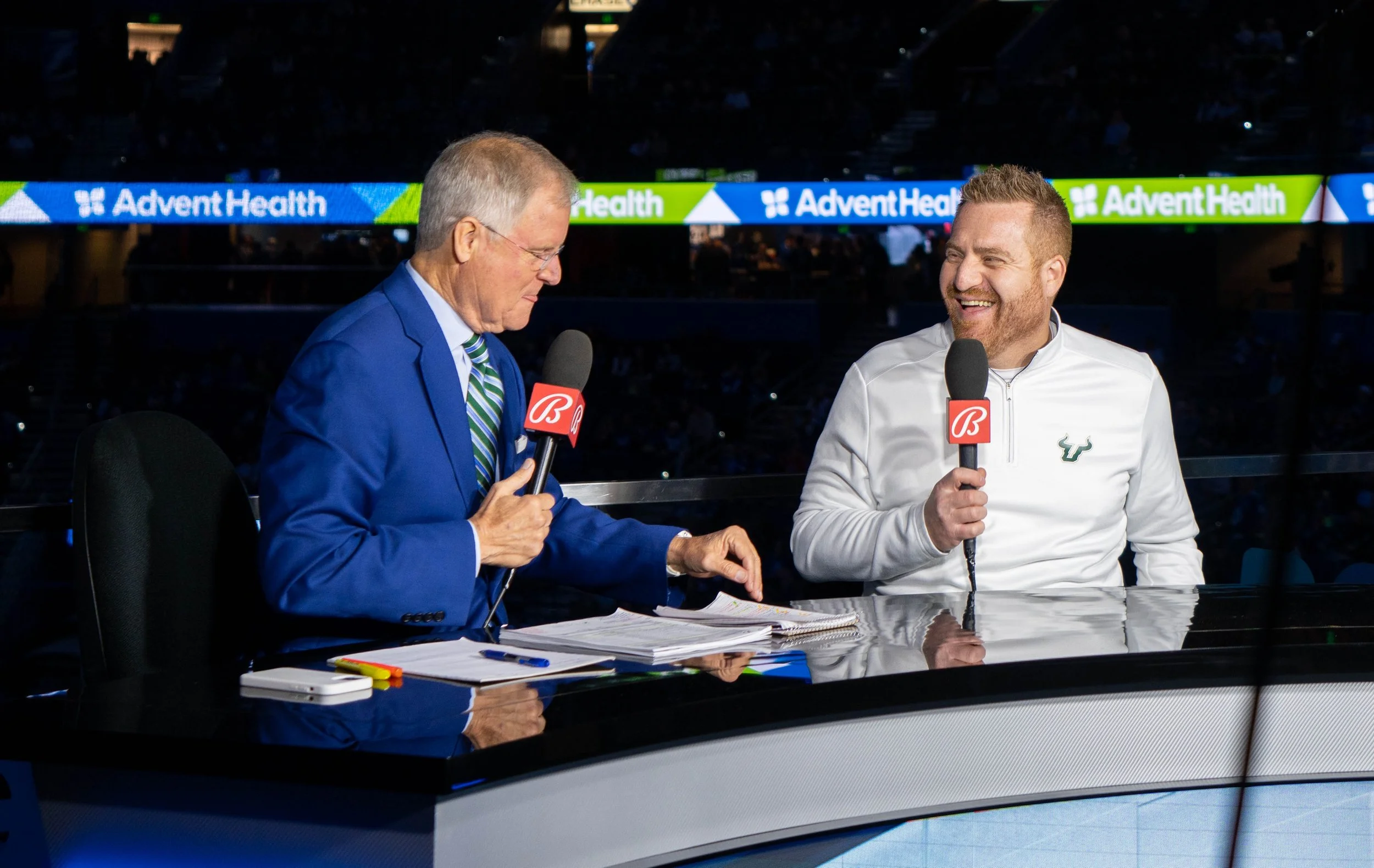 Two men sitting at a broadcast desk, holding microphones, engaging in a conversation during a sports event. The man on the left wears a blue suit and glasses, while the man on the right wears a white jacket with a sports logo.