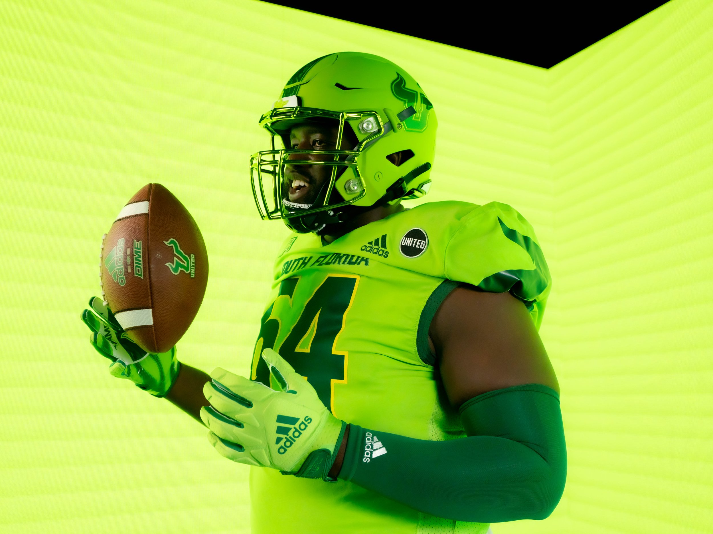 American football player in bright yellow uniform holding a football, wearing a helmet and gloves, posing against a yellow background.
