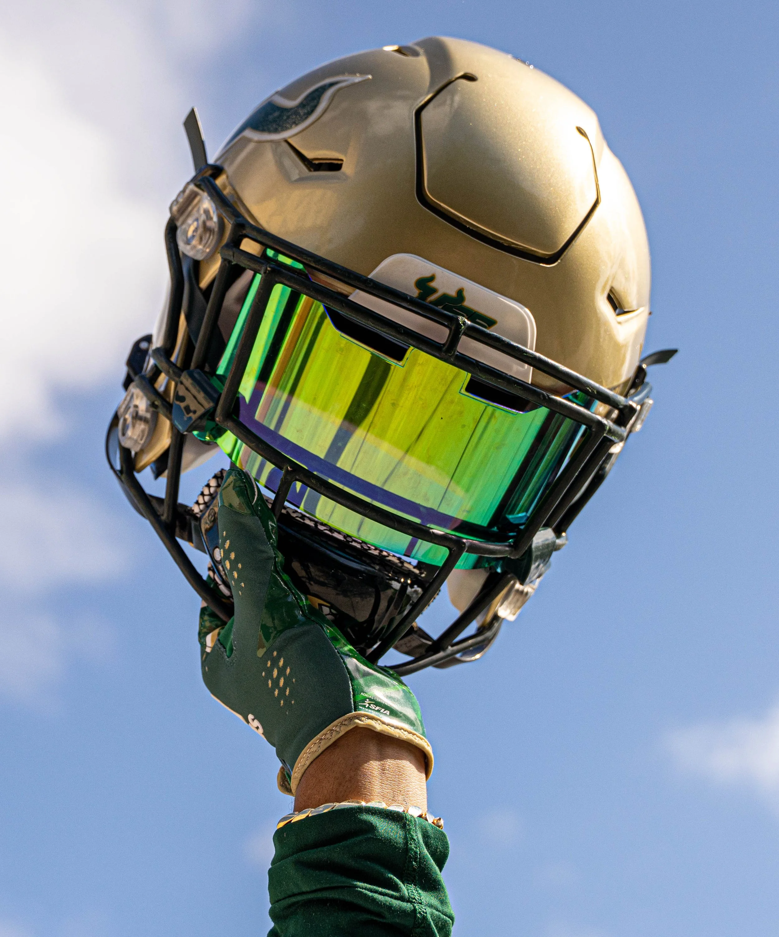 A football helmet with a reflective visor being held up by a person wearing a green glove against a blue sky background.