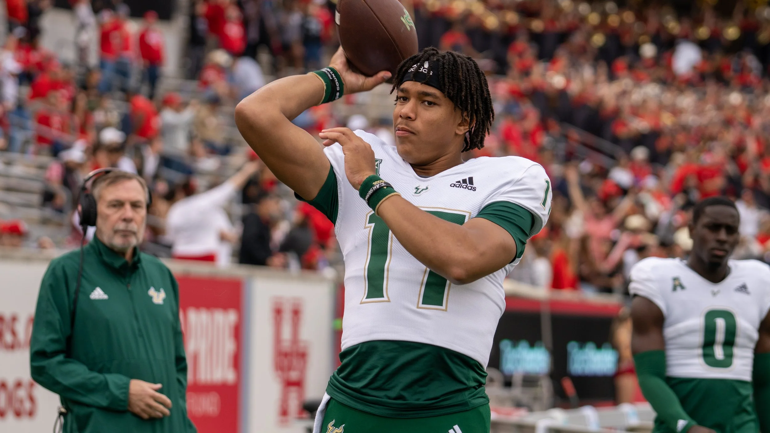 A football player with jersey number 17 in a green and white uniform about to throw a football during a game, with fans in the background.